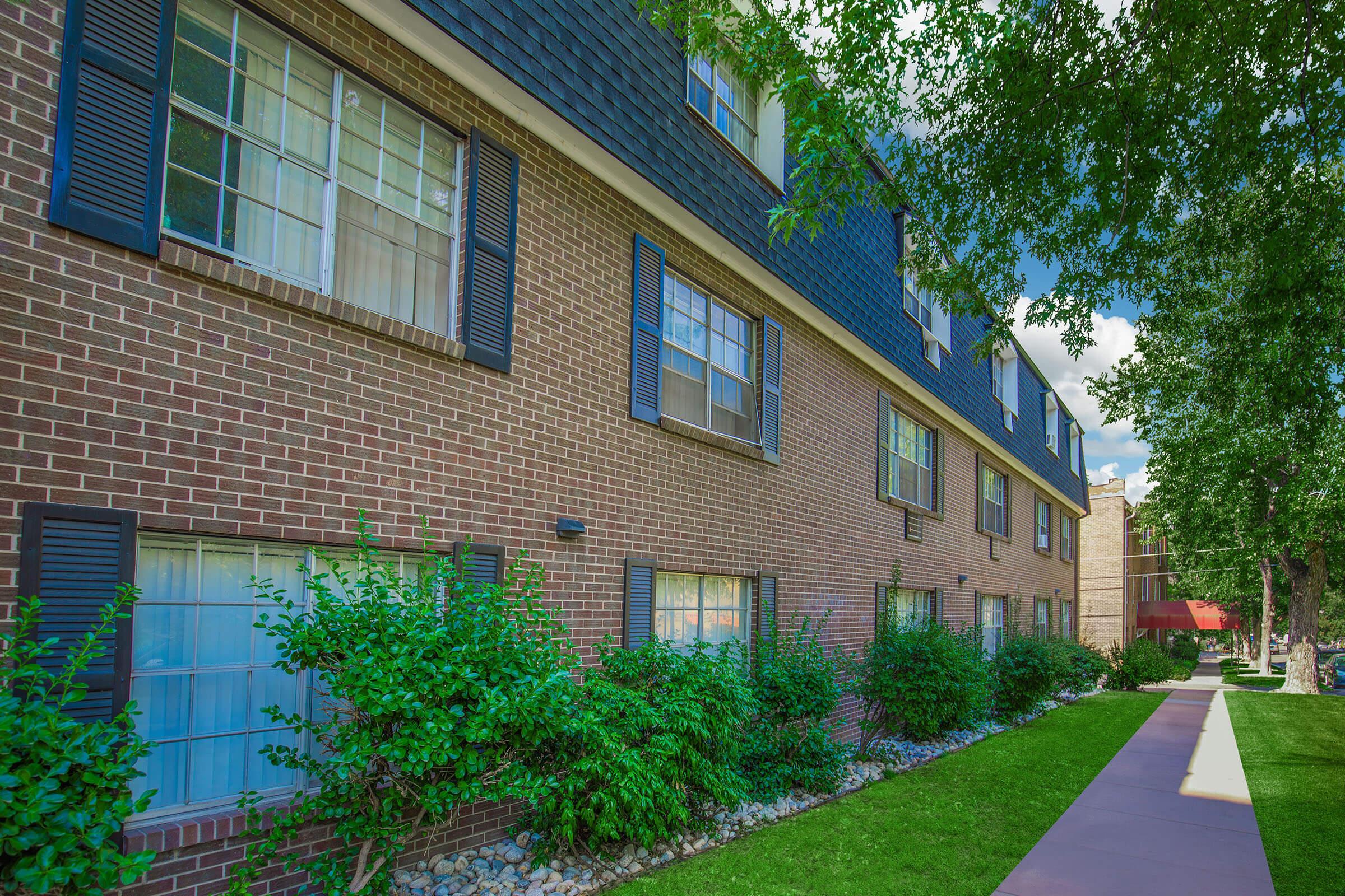 A two-story brick building with multiple windows, each featuring black shutters. The exterior is surrounded by manicured greenery and a path leading to the entrance. Bright sunlight illuminates the scene, creating a vibrant atmosphere with blue skies and scattered clouds.