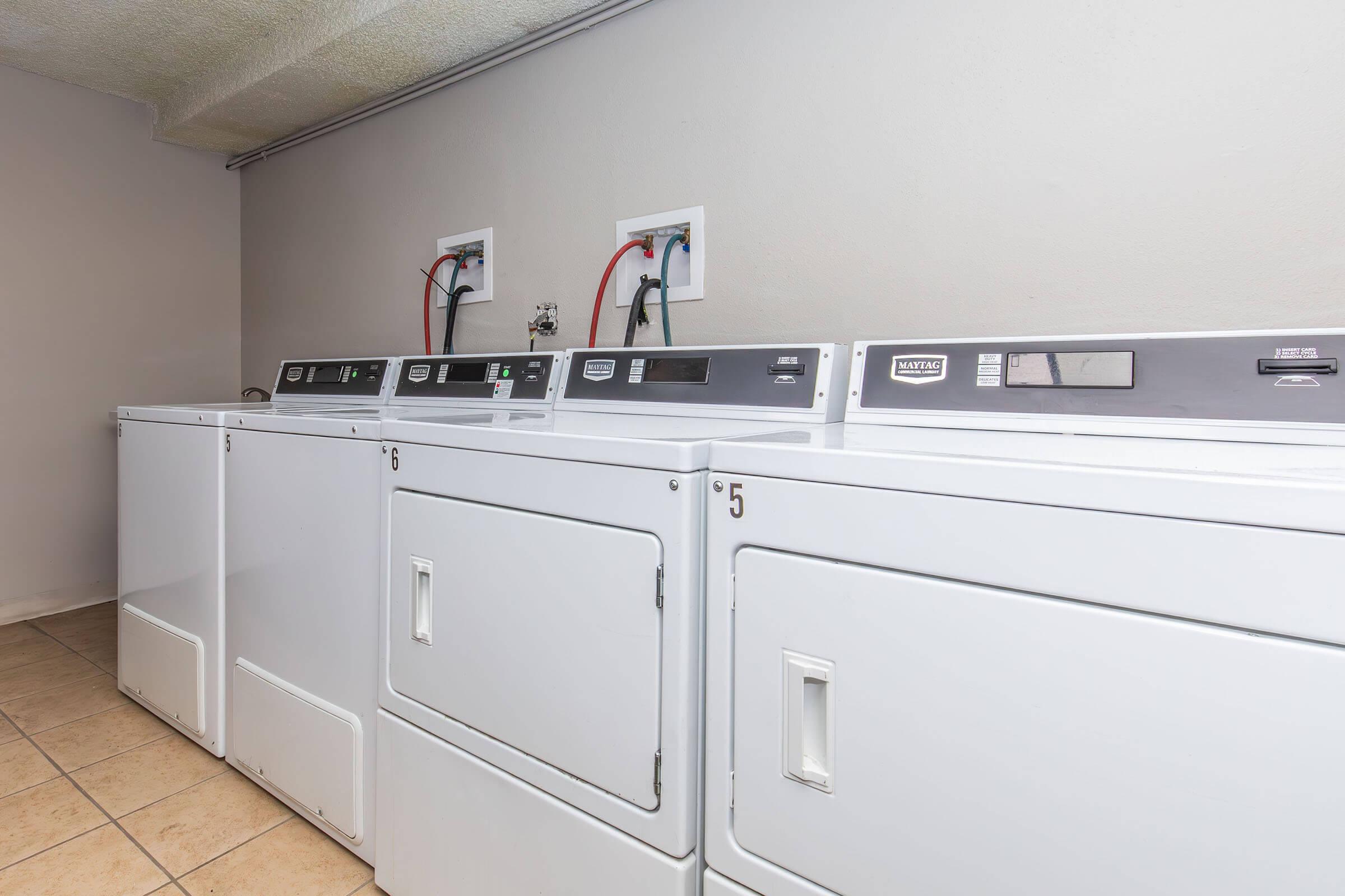 A row of white washing machines and dryers in a clean, well-lit laundry room. The machines are lined up against a beige wall, with utility connections visible above them. The floor is tiled, and the overall space appears organized and functional.