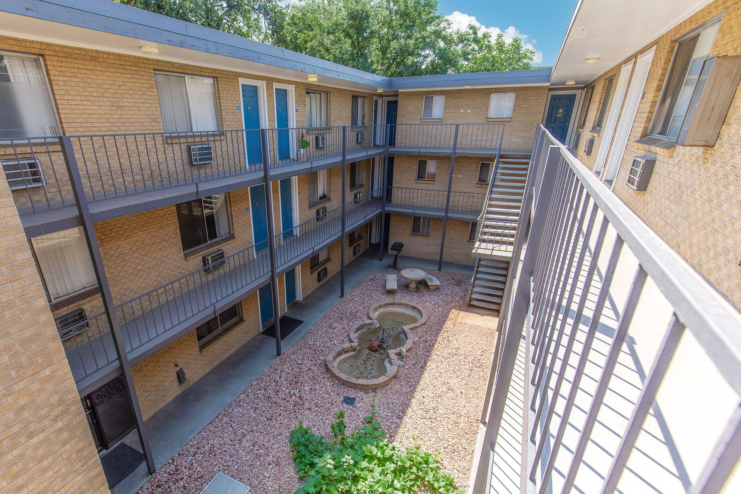 View of an apartment complex courtyard featuring a central fountain surrounded by gravel and greenery. The building consists of multiple stories with balconies and individual entryways visible. Bright daylight illuminates the scene, highlighting the brick exterior and open design.