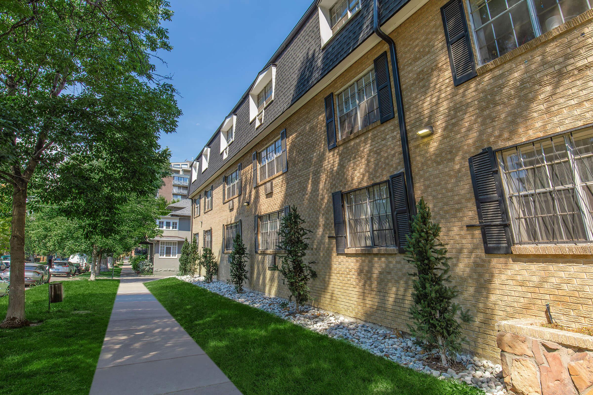 A sunny street view of a residential building with a brick exterior. The facade features several windows with shutters and is bordered by neatly trimmed grass and white stones. Trees provide shade along the sidewalk, creating a pleasant atmosphere in a suburban setting.