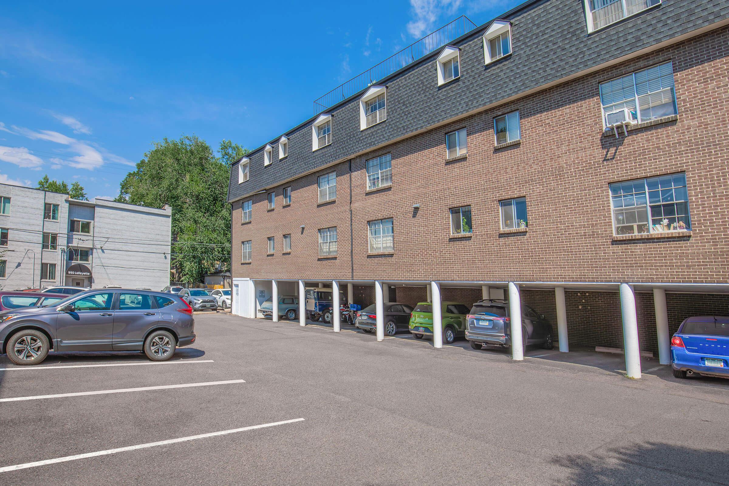 A multi-story brick apartment building with several windows and a flat roof. The foreground features a parking area with multiple vehicles, some parked under covered spaces. The sky is clear with a few clouds, and there are trees in the background.