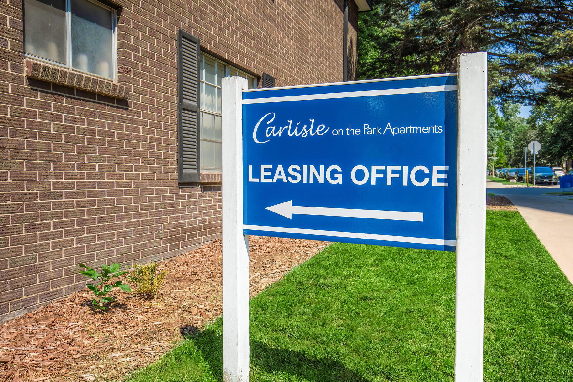 Blue sign indicating the leasing office for Carlisle on the Park Apartments, with an arrow pointing to the left. The sign is set against a brown brick building and green grass, surrounded by landscaping.