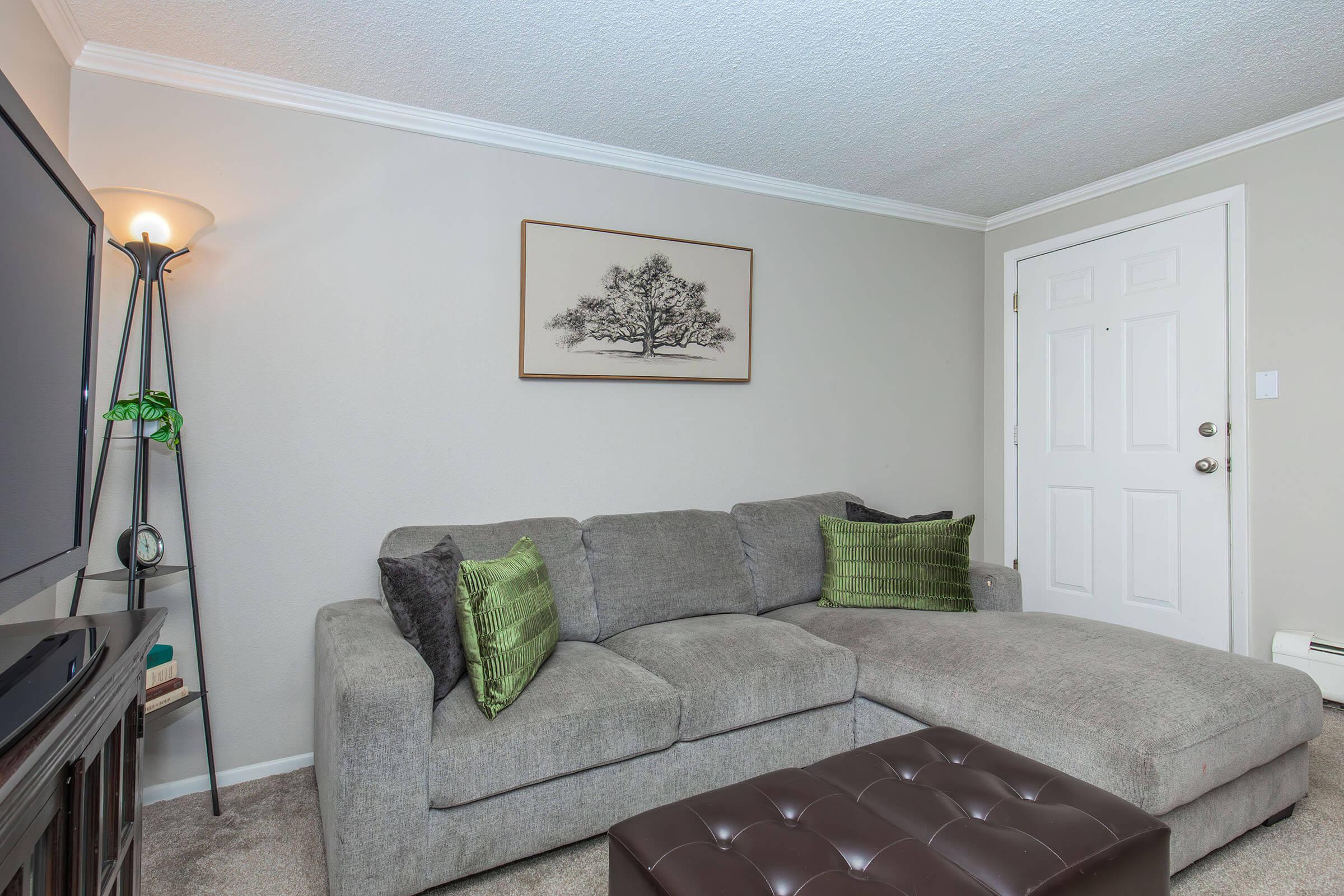 A cozy living room featuring a gray sectional sofa with green decorative pillows, a brown tufted ottoman, and a standing lamp. There is a wall-mounted art piece depicting a tree. The room is well-lit with a light-colored wall and has a door visible in the background.