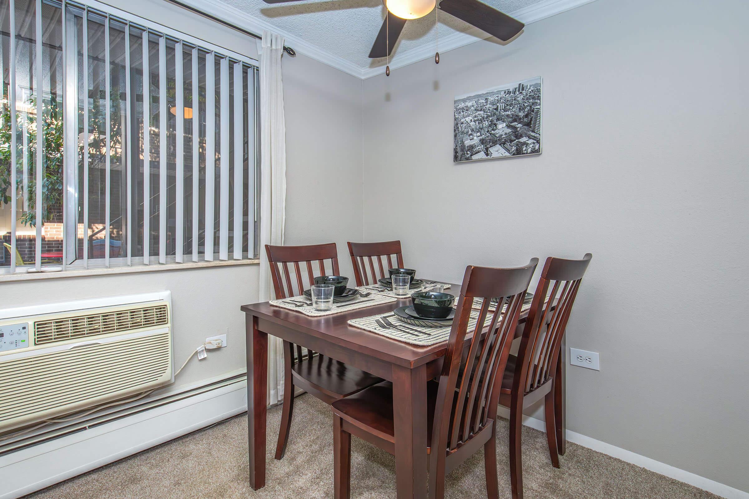 A cozy dining area featuring a wooden table set for four, with black bowls and placemats. A ceiling fan is overhead, and a window with blinds allows natural light. An air conditioning unit is visible on one wall, and a black-and-white photograph hangs above the table.