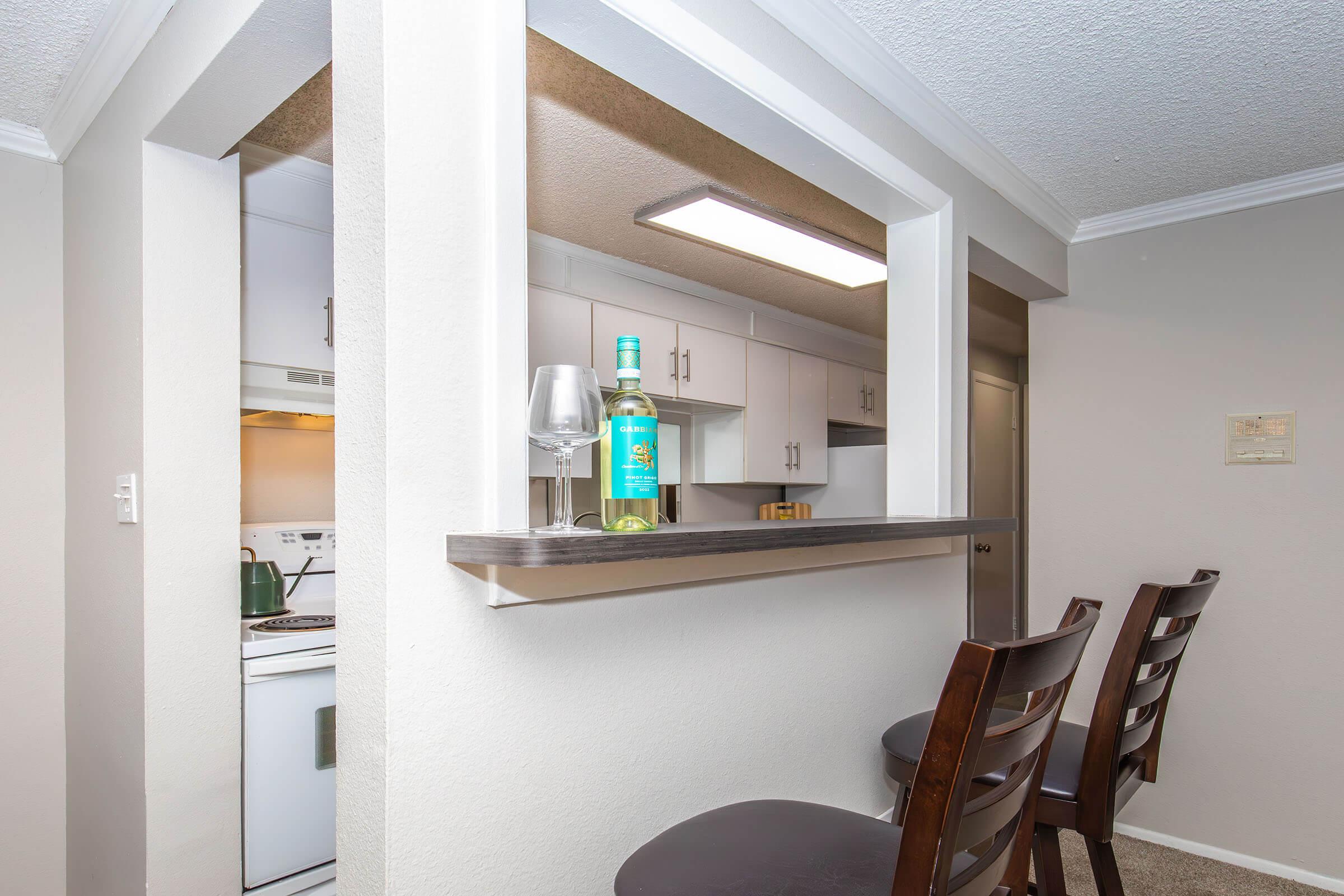 A cozy kitchen area featuring a bar with two wooden stools. A bottle of white wine and a wine glass sit on the bar, overlooking a well-equipped kitchen with white cabinetry and modern appliances. The walls are painted light beige, creating a warm and inviting atmosphere.