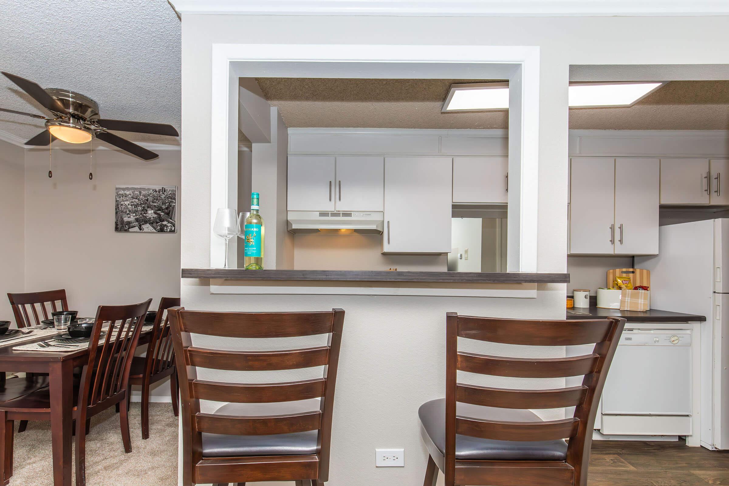 A view from a modern kitchen with a breakfast bar, featuring two wooden stools. In the background, there's a dining area with a table set for meals. The kitchen is equipped with white cabinets and appliances, and a glass of wine and a bottle sit on the counter, adding to a cozy atmosphere.