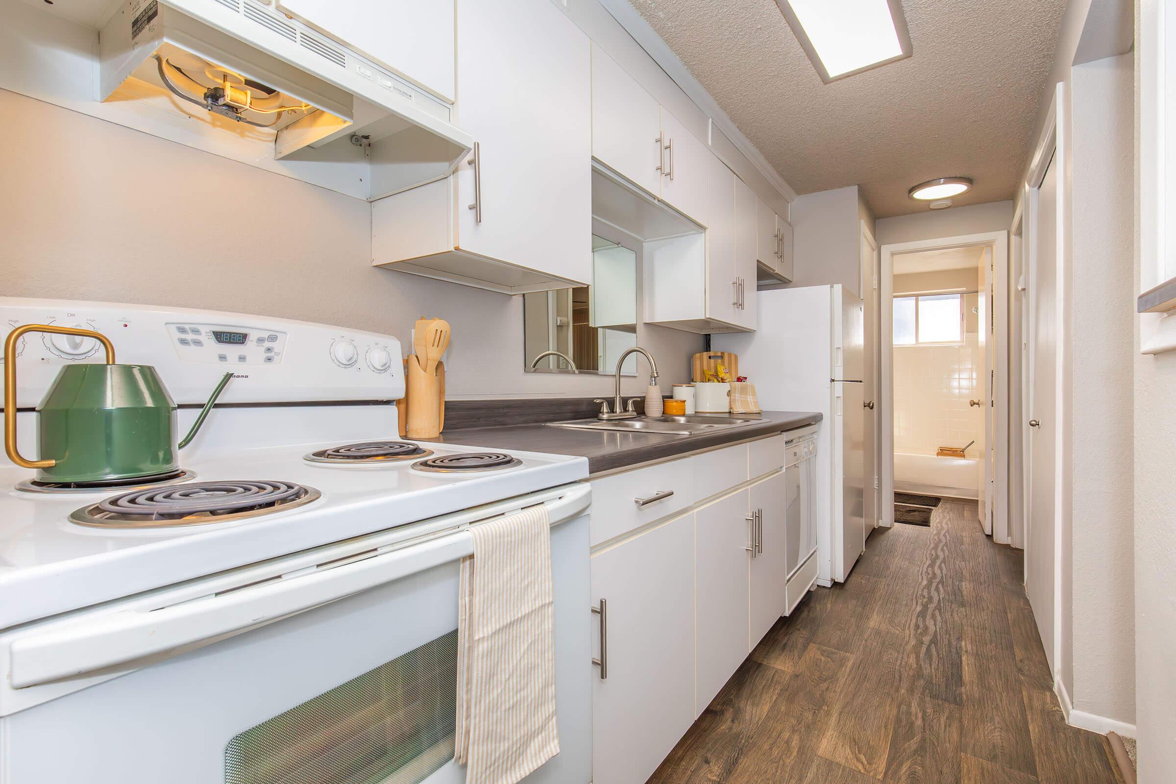 A bright, modern kitchen featuring white cabinets and appliances, a stove with a hood, a sink, and wooden utensils. The hallway leads to a bathroom with light-colored tiles, adding to the clean and spacious atmosphere. Natural light illuminates the area, enhancing the inviting feel of the space.