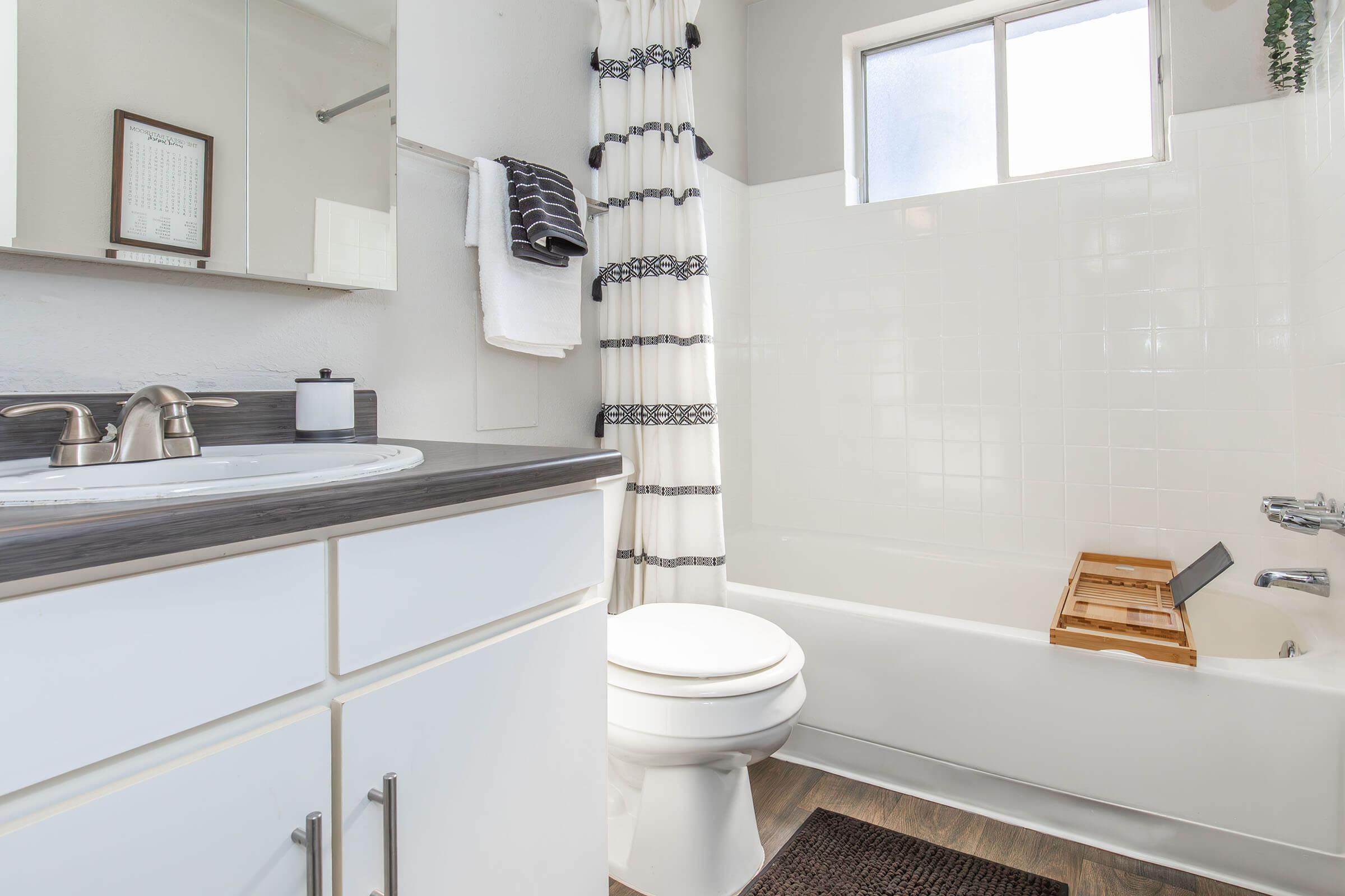 A modern bathroom featuring a white bathtub with a wooden bath tray, a white toilet, a sink with a dark countertop, and a large mirror. There are towels hanging on a rack and a shower curtain in a light color with decorative stripes. Natural light filters in through a window.