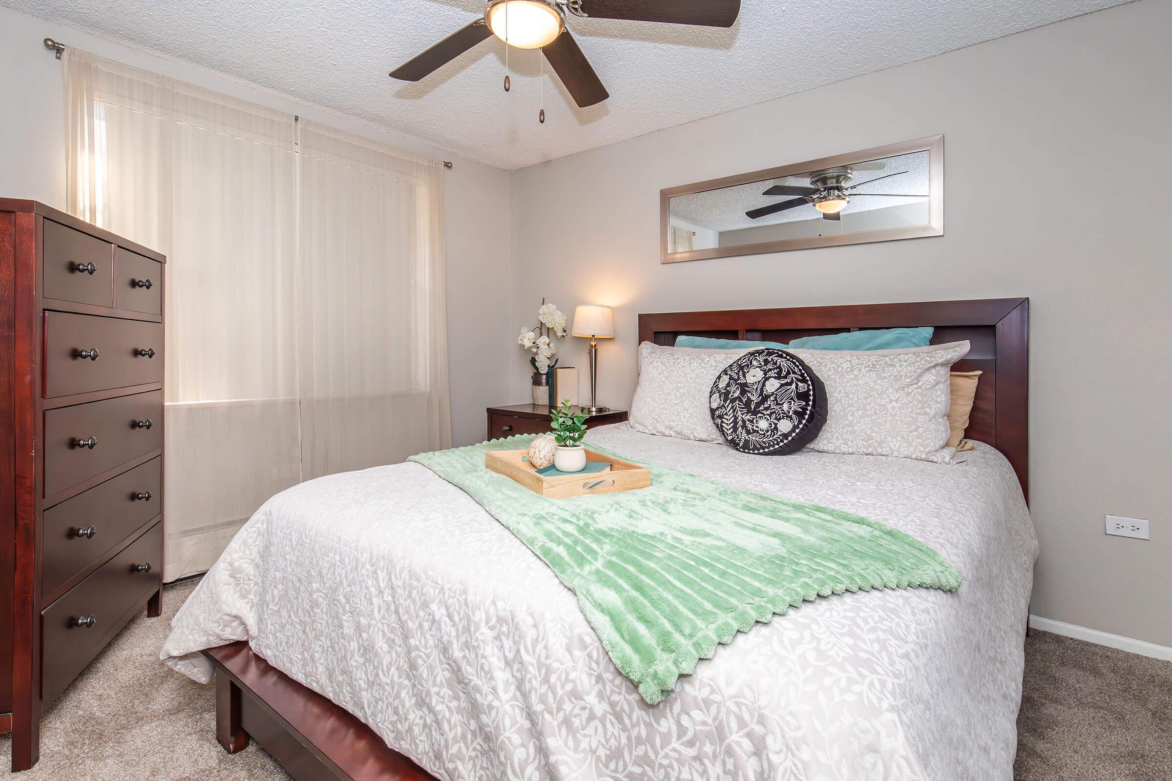 A cozy bedroom featuring a queen-sized bed with decorative pillows and a soft green throw. There is a wooden nightstand with a lamp, a small tray on the bed, and a chest of drawers. A large mirror and sheer curtains enhance the natural light in the room, creating a warm and inviting atmosphere.