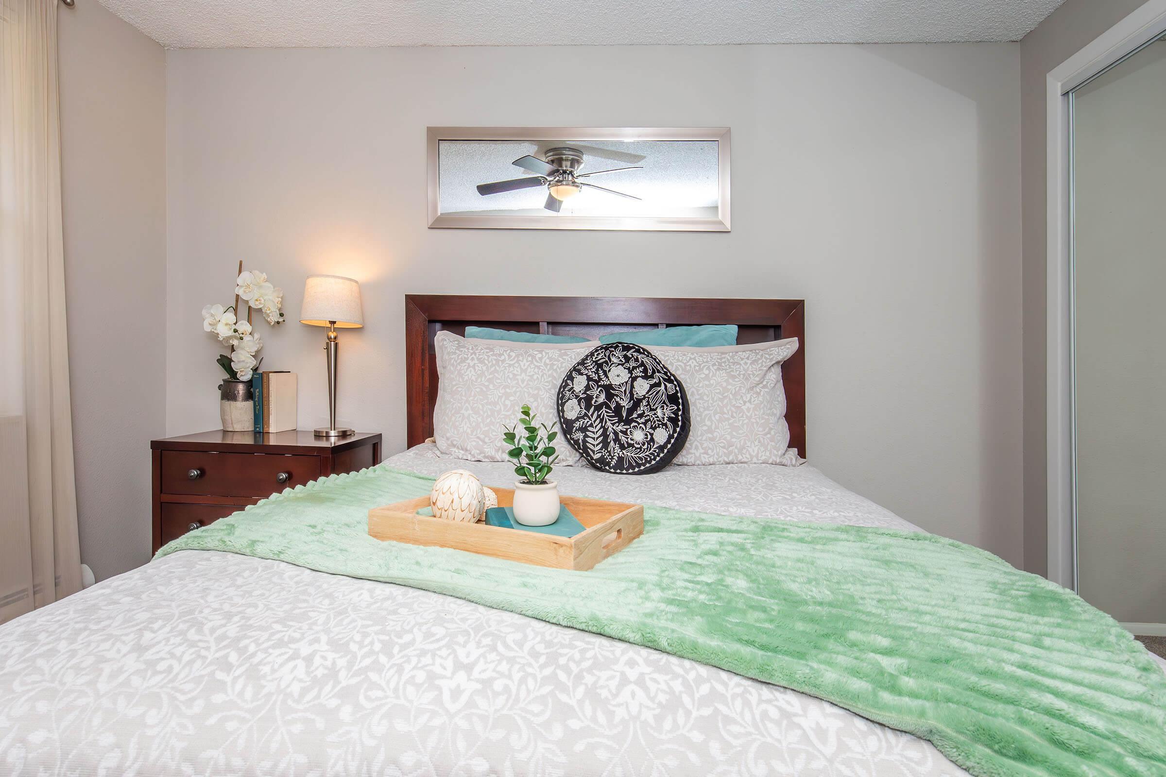 A cozy bedroom featuring a neatly made bed with a light green throw blanket and decorative pillows. A wooden nightstand with a lamp and a small bouquet of flowers sits beside the bed, while a tray with a decorative orb and a small plant rests on the bed. A mirror and a closet door are visible in the background.