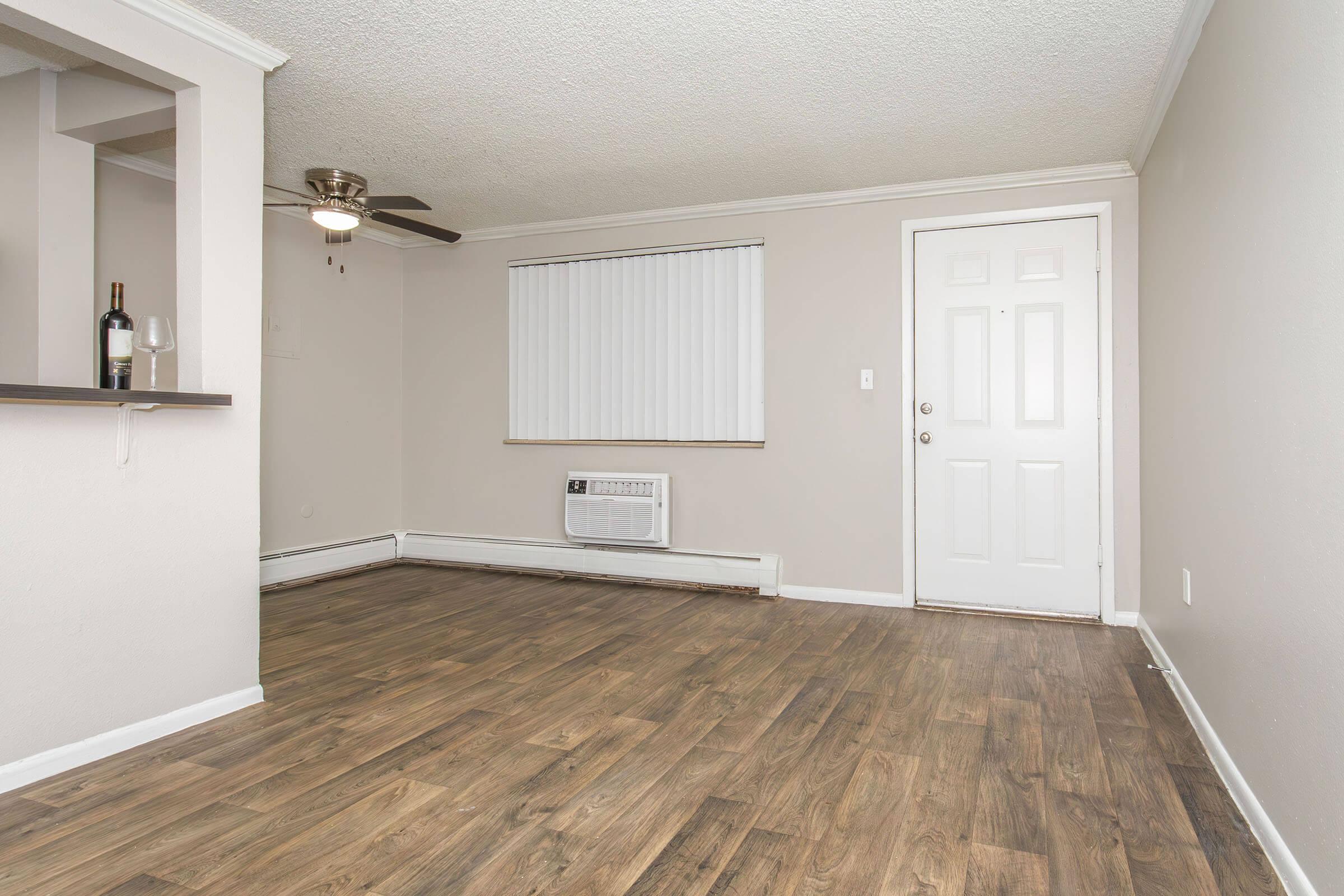 Interior view of a vacant living room featuring light-colored walls, a fan on the ceiling, and a door leading outside. There is an air conditioning unit on the floor, a wine bottle and glass on a small shelf, and wooden flooring. A window with vertical blinds allows natural light in.