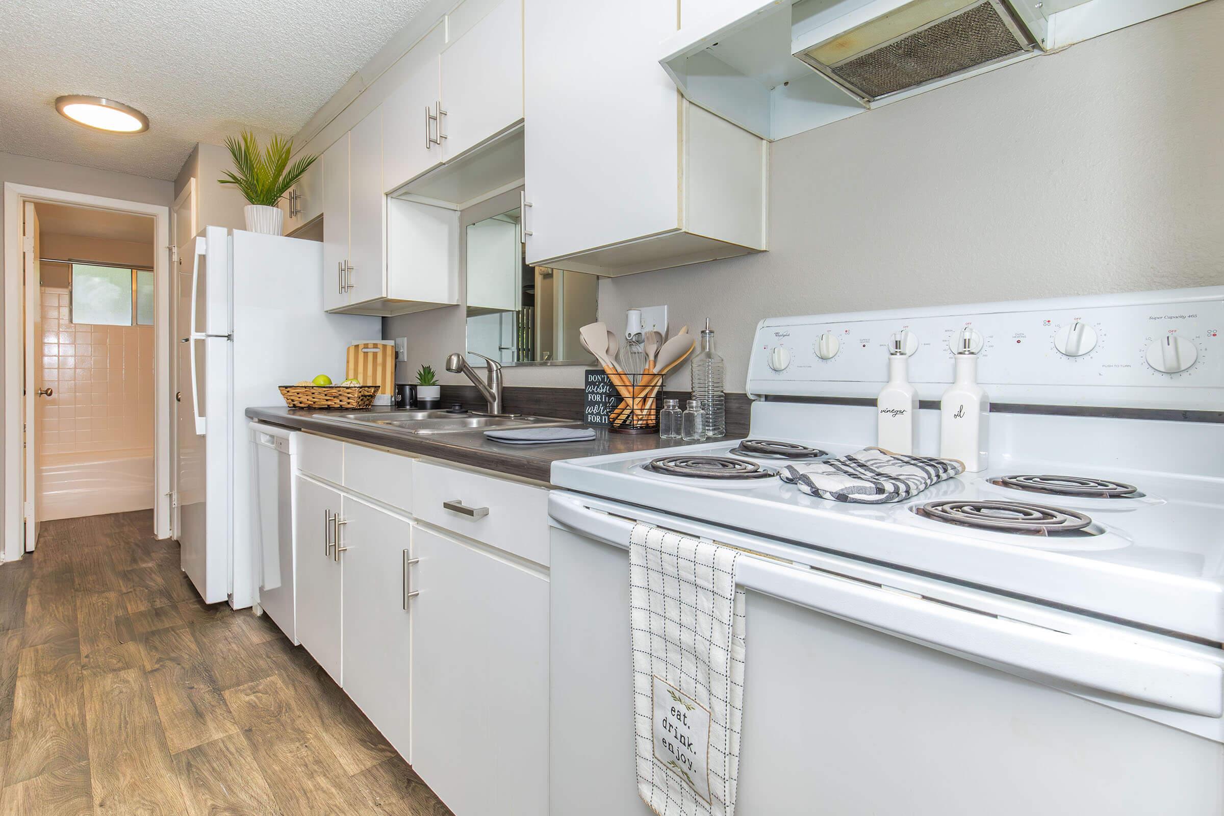 A modern kitchen featuring white cabinets, a gray countertop, and an electric stove. The setup includes a refrigerator, kitchen utensils, and decorative plants. Brightly lit with natural light, the space has a clean and minimalist design, complemented by a towel hanging on the oven.
