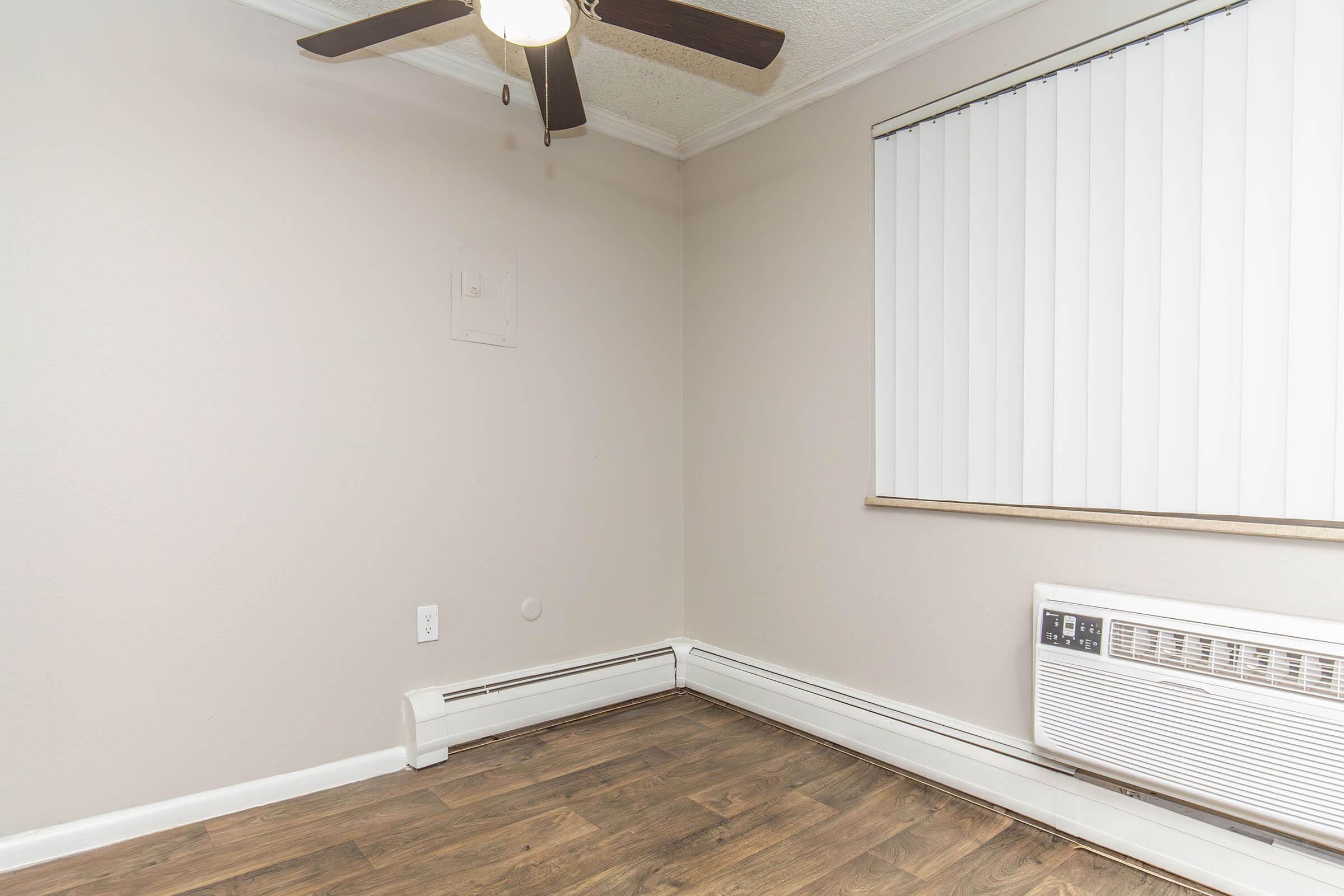 Interior view of a small room featuring a ceiling fan, a wall-mounted air conditioning unit, and a window with vertical blinds. The walls are painted light beige, and the flooring appears to be dark wood laminate. The corner of the room is mostly empty.