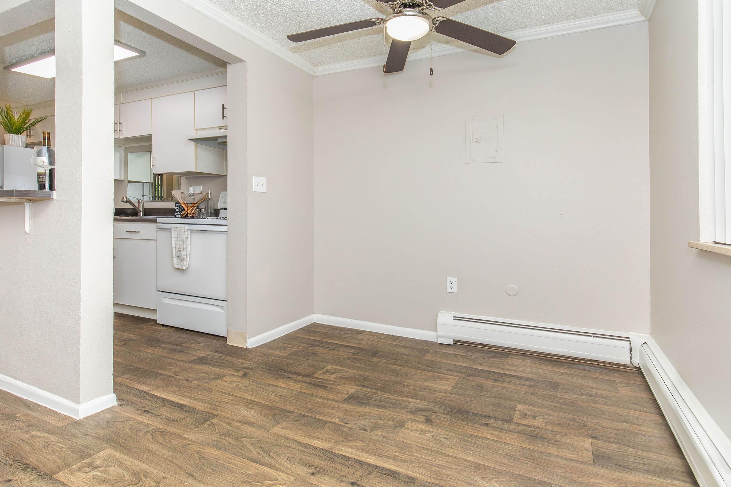 A well-lit apartment interior featuring a ceiling fan, beige walls, and wooden flooring. There is a kitchenette with white appliances visible in the background, and a small shelf displaying decorative items on the left. A large window allows natural light to enter the space, creating a cozy atmosphere.