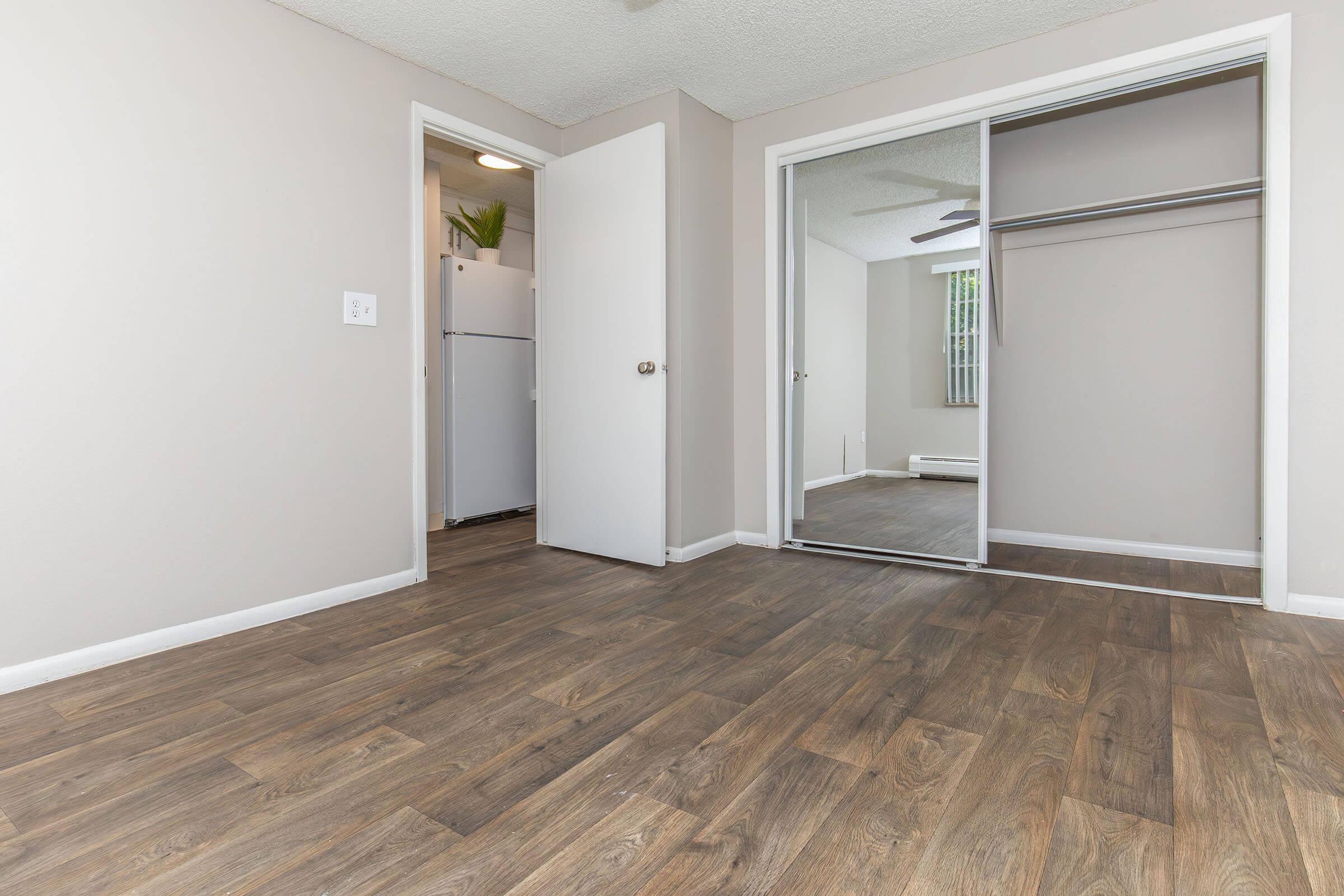 A vacant room featuring a light-colored wall, a mirrored closet door, and wooden floors. The room has a doorway leading to another area, and a door partially open revealing an adjacent space. Natural light filters in through a window, creating a bright and airy atmosphere.
