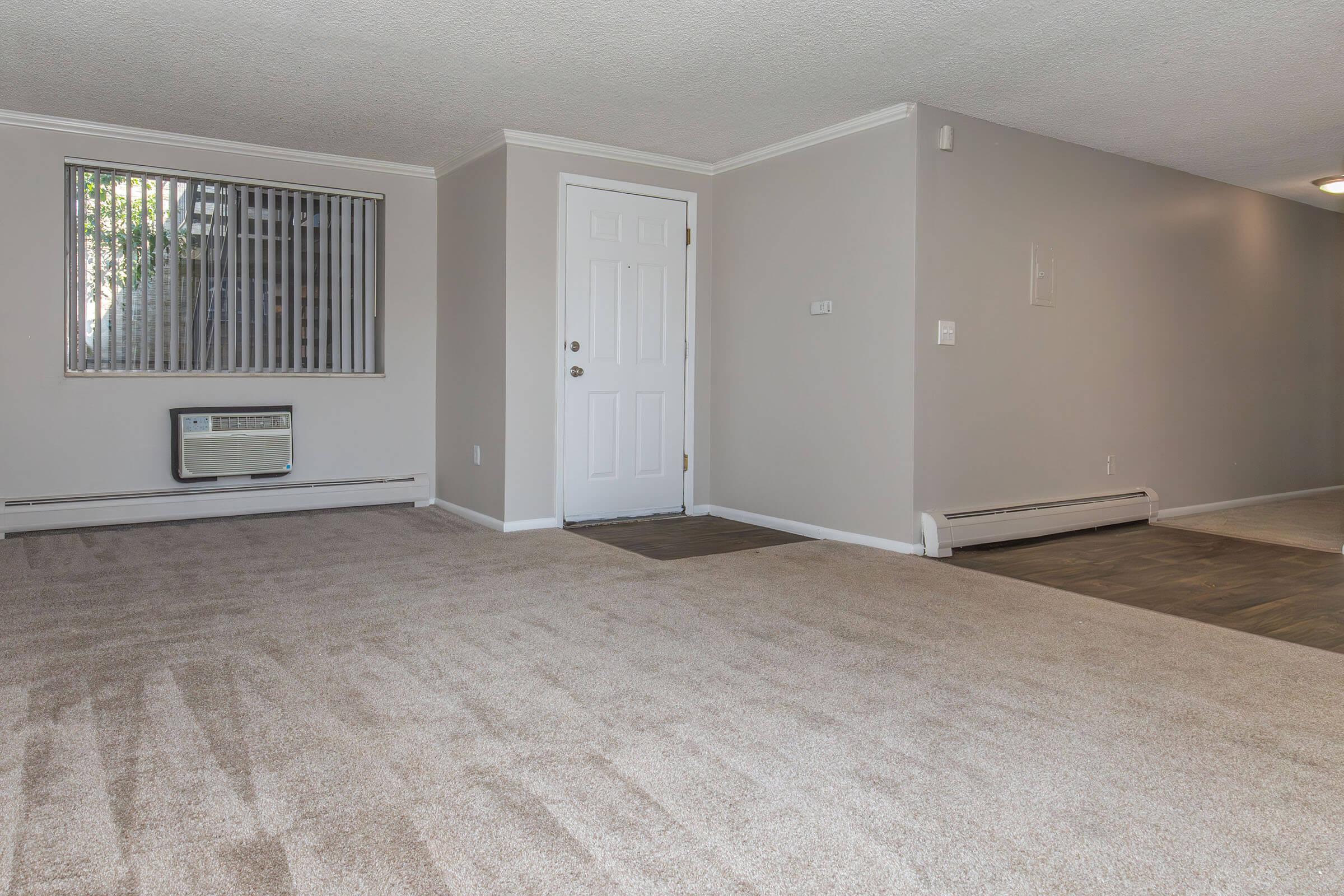 A spacious, carpeted living area featuring an air conditioning unit near a large window with vertical blinds. The walls are painted in a light color, and there is a front door on the right. The room has a clean, inviting appearance with natural light coming in.