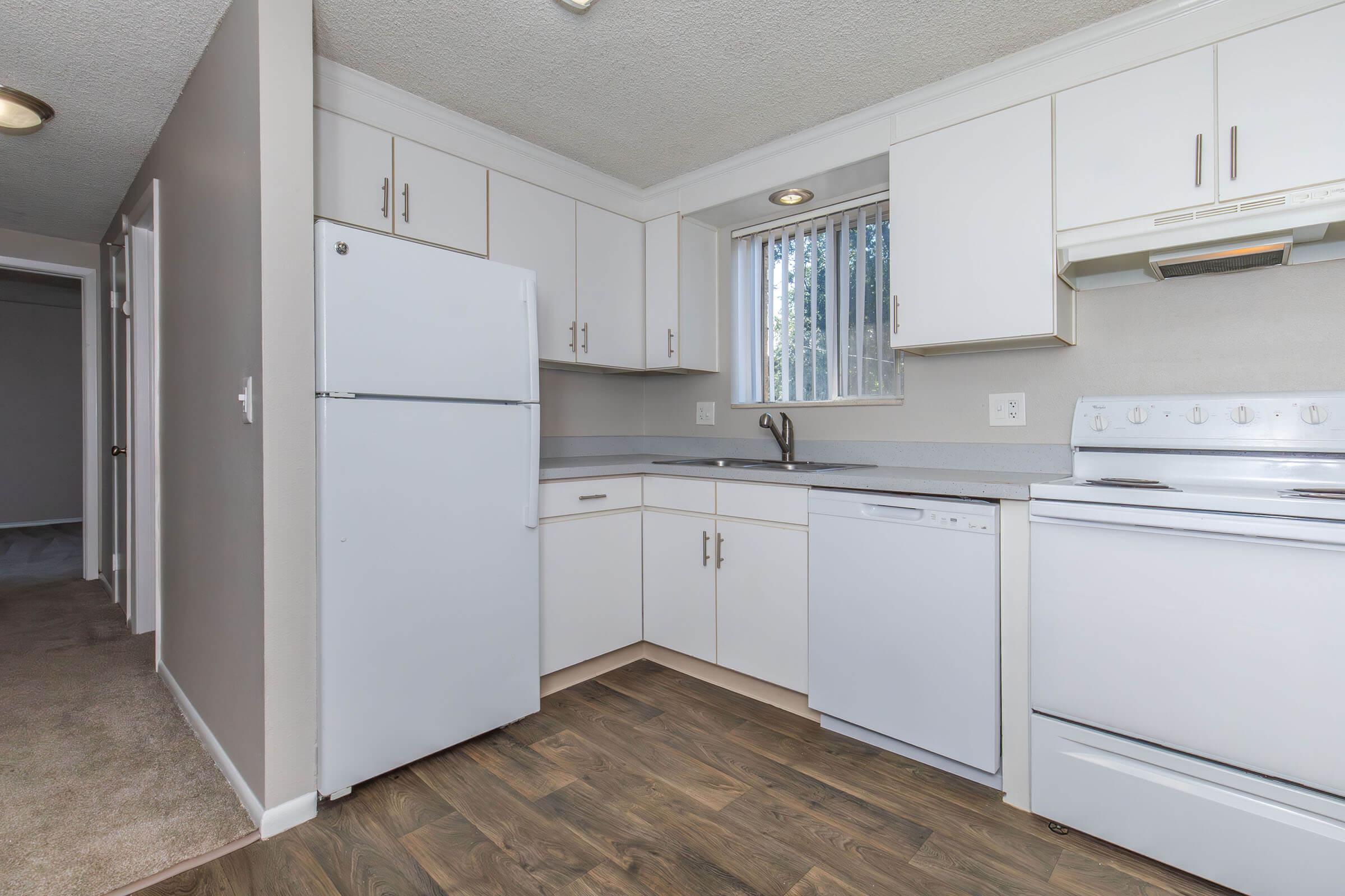 A modern kitchen featuring white cabinetry, a refrigerator, stove, and dishwasher. The countertop is light gray, and there is a window for natural light. The floor is wood-like, and the walls are painted in neutral tones. There is a hallway visible in the background.