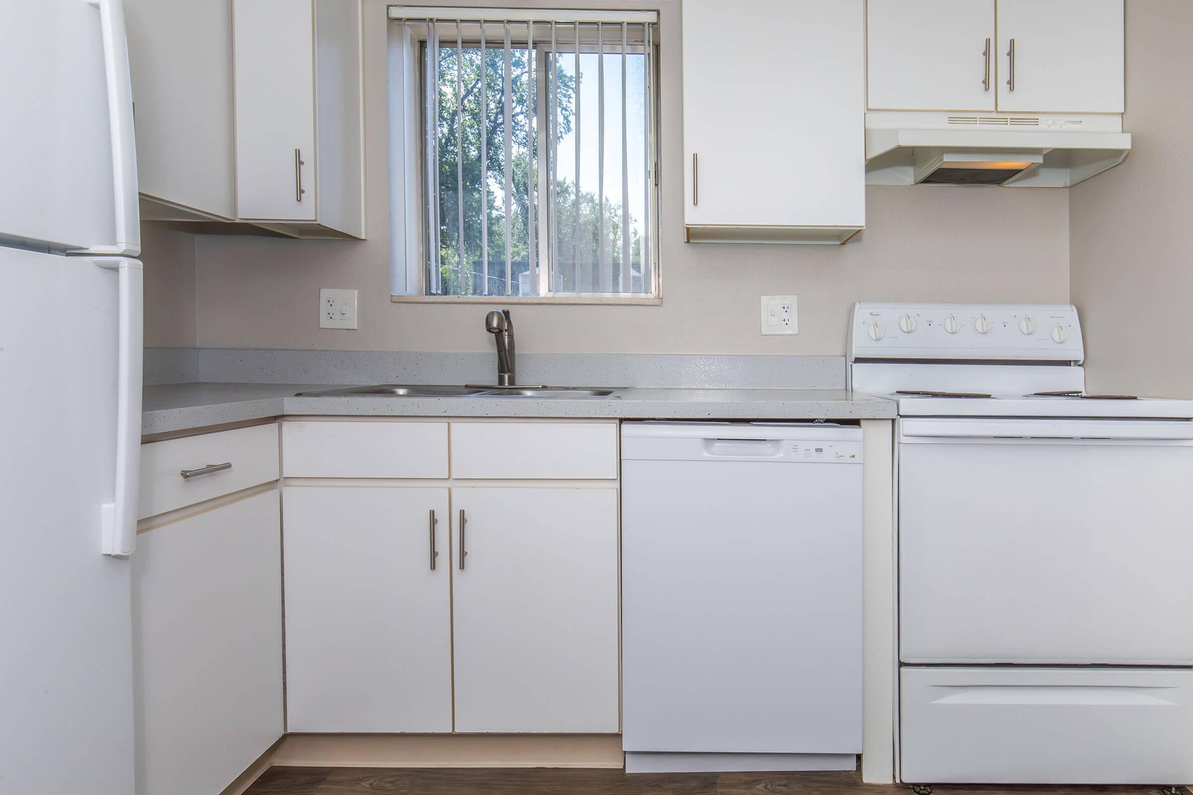 A modern kitchen featuring white cabinetry, a sink beneath a window with vertical blinds, a refrigerator, a dishwasher, and a stovetop with oven. The countertop is light-colored, and the flooring is wooden. Natural light comes through the window, creating a bright atmosphere.