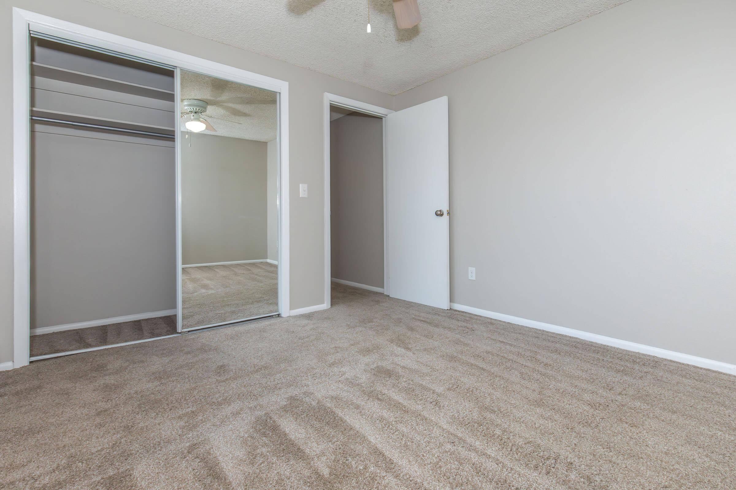 A clean, empty room with light beige carpet and neutral gray walls. A mirrored closet door reflects the space, and a doorway leads to another room. A ceiling fan is visible, adding to the bright, airy atmosphere.