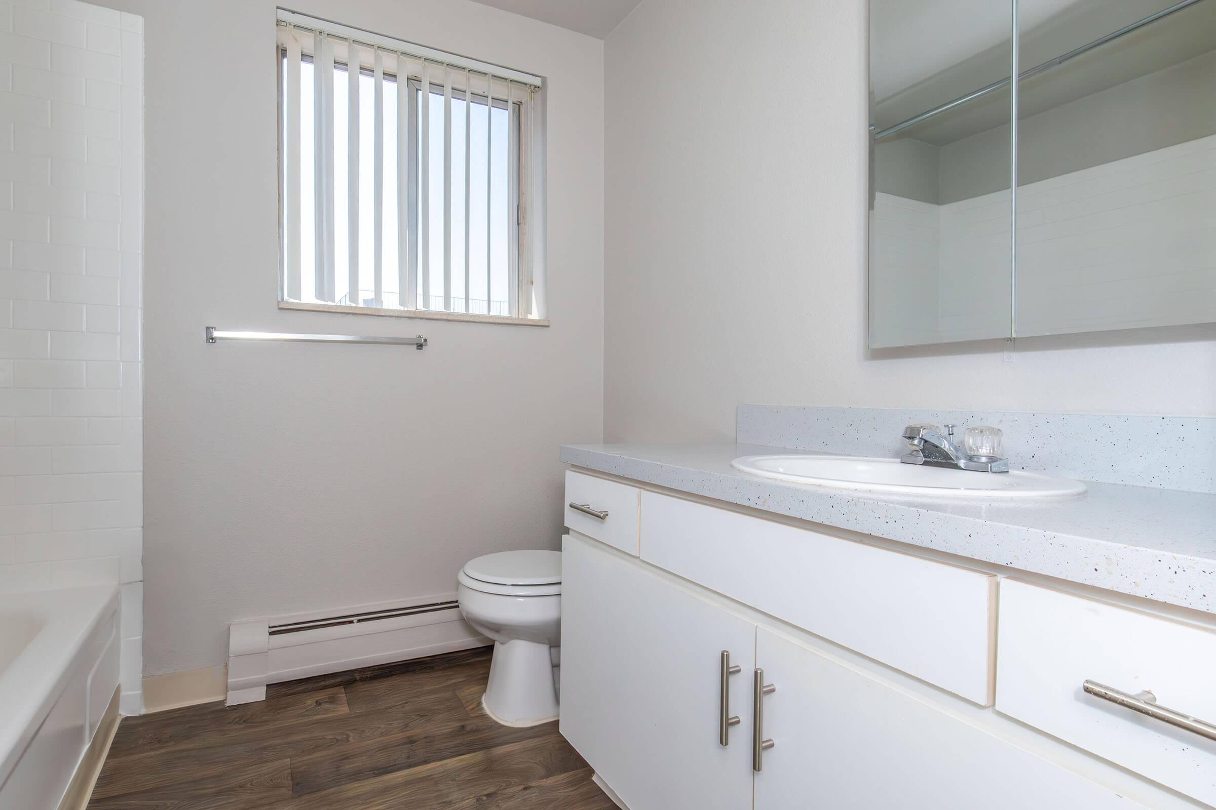 Bright and modern bathroom featuring a white sink and countertop, a toilet, a bathtub, and a large mirror. Natural light enters through a window with vertical blinds. The flooring is a wood-like laminate, enhancing the clean and contemporary look of the space.