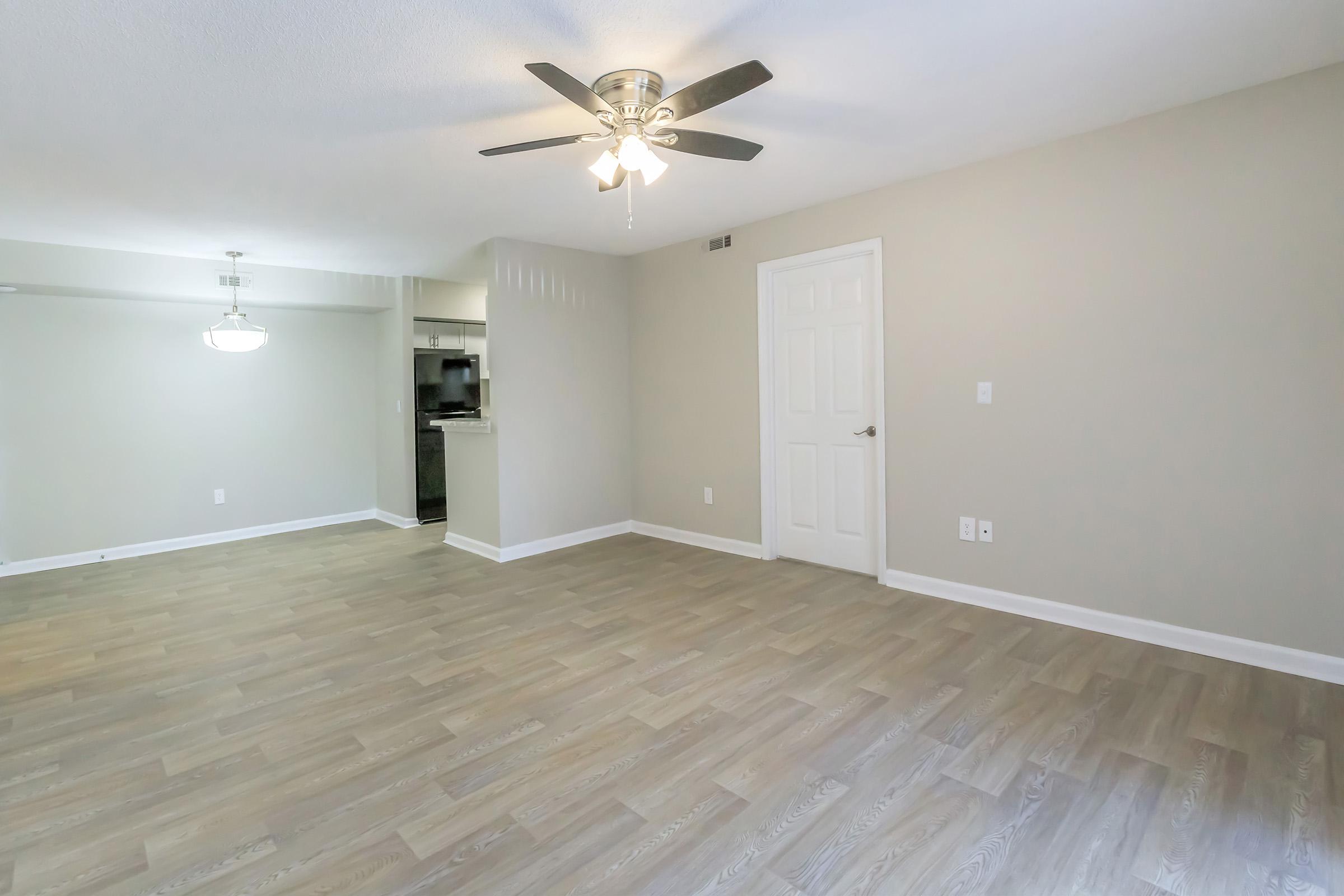 A spacious, light-filled room with light wood flooring and a ceiling fan. The walls are painted a soft neutral color, and there is a doorway leading to a kitchen area in the background. The room is empty, creating a sense of openness and possibilities for interior design.