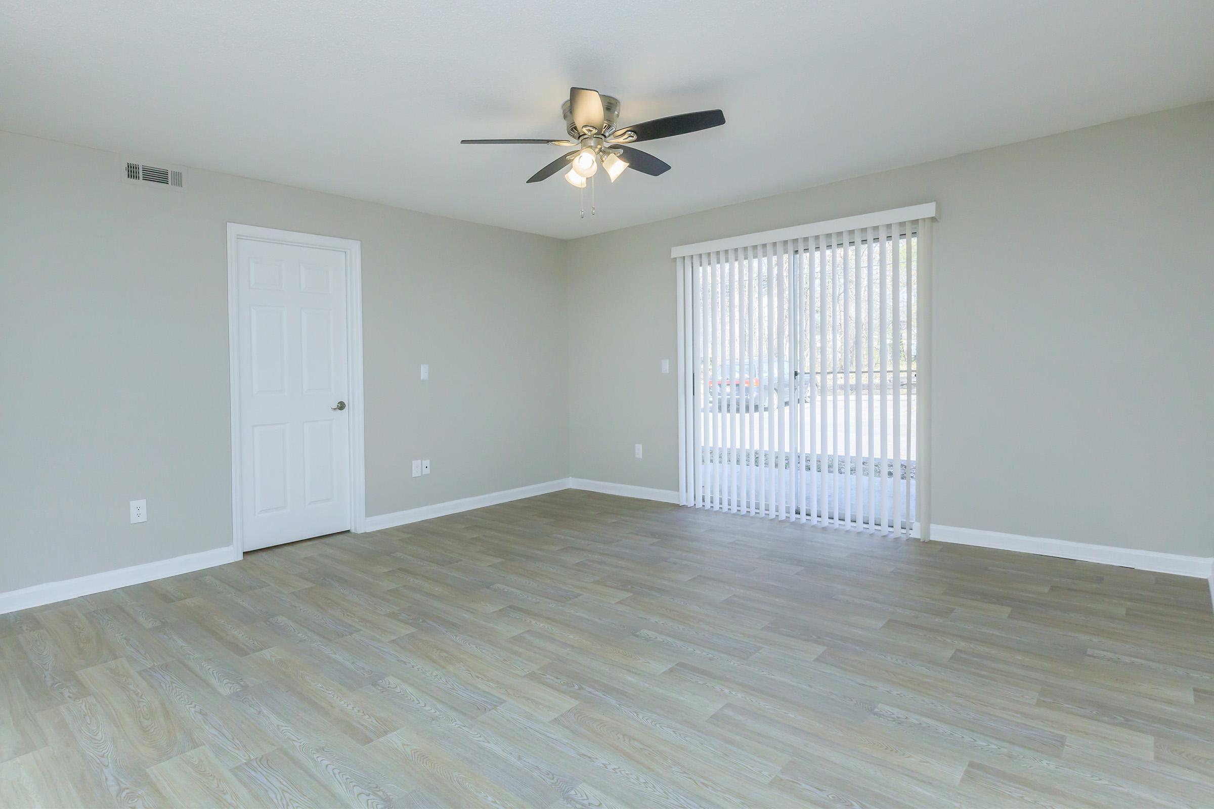 A spacious, empty living room featuring light gray walls, a ceiling fan, and large windows with vertical blinds. The flooring is a light wood laminate, creating a warm ambiance. A door is visible on the left side, and the overall space is bright and airy.