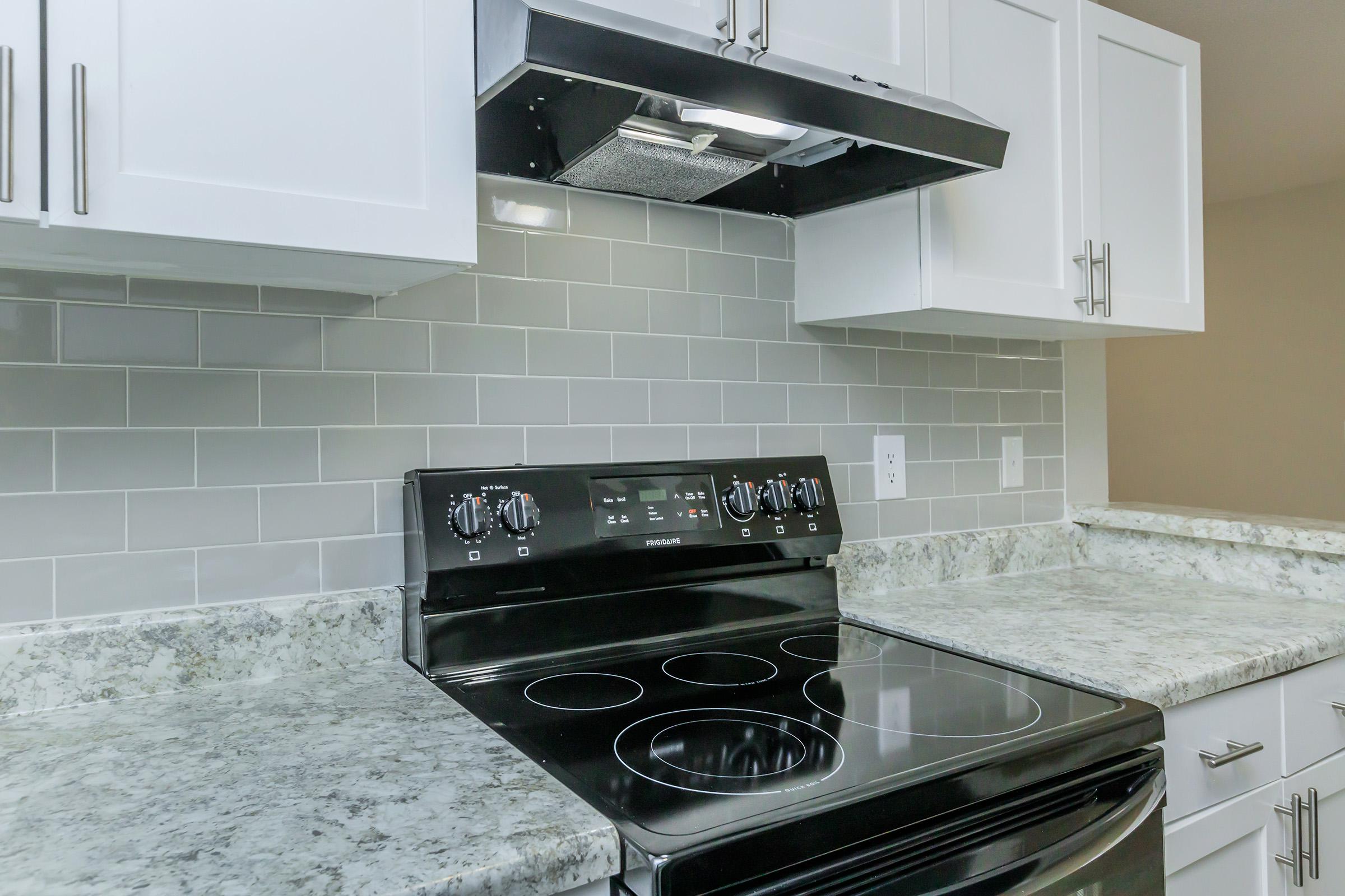 A modern kitchen featuring a black electric stove with control knobs and a built-in range hood, set against a white cabinetry backdrop. The countertops are made of light-colored granite, and the backsplash consists of sleek gray tiles, creating a contemporary and stylish cooking space.