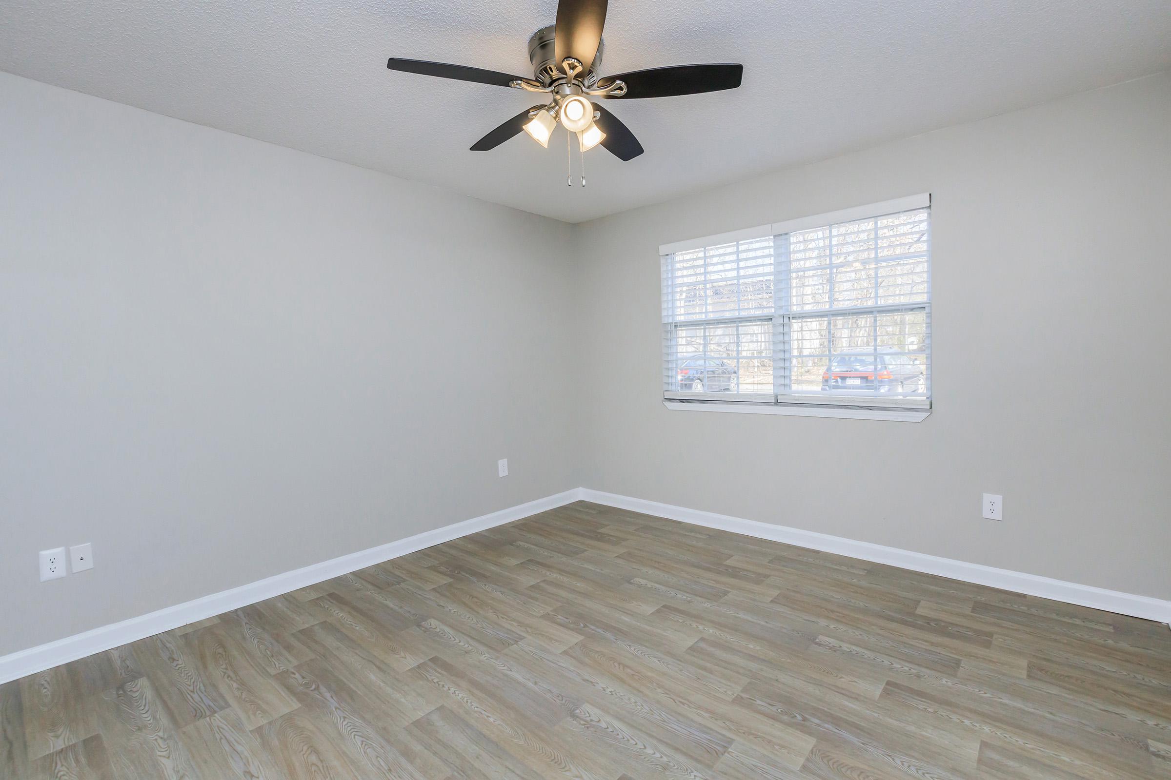 A well-lit, empty room featuring light gray walls, a ceiling fan with three bulbs, and a large window with blinds. The floor has a wood-like design, and there's space for furniture. The room appears freshly painted and maintained, ideal for various uses like a bedroom or office.