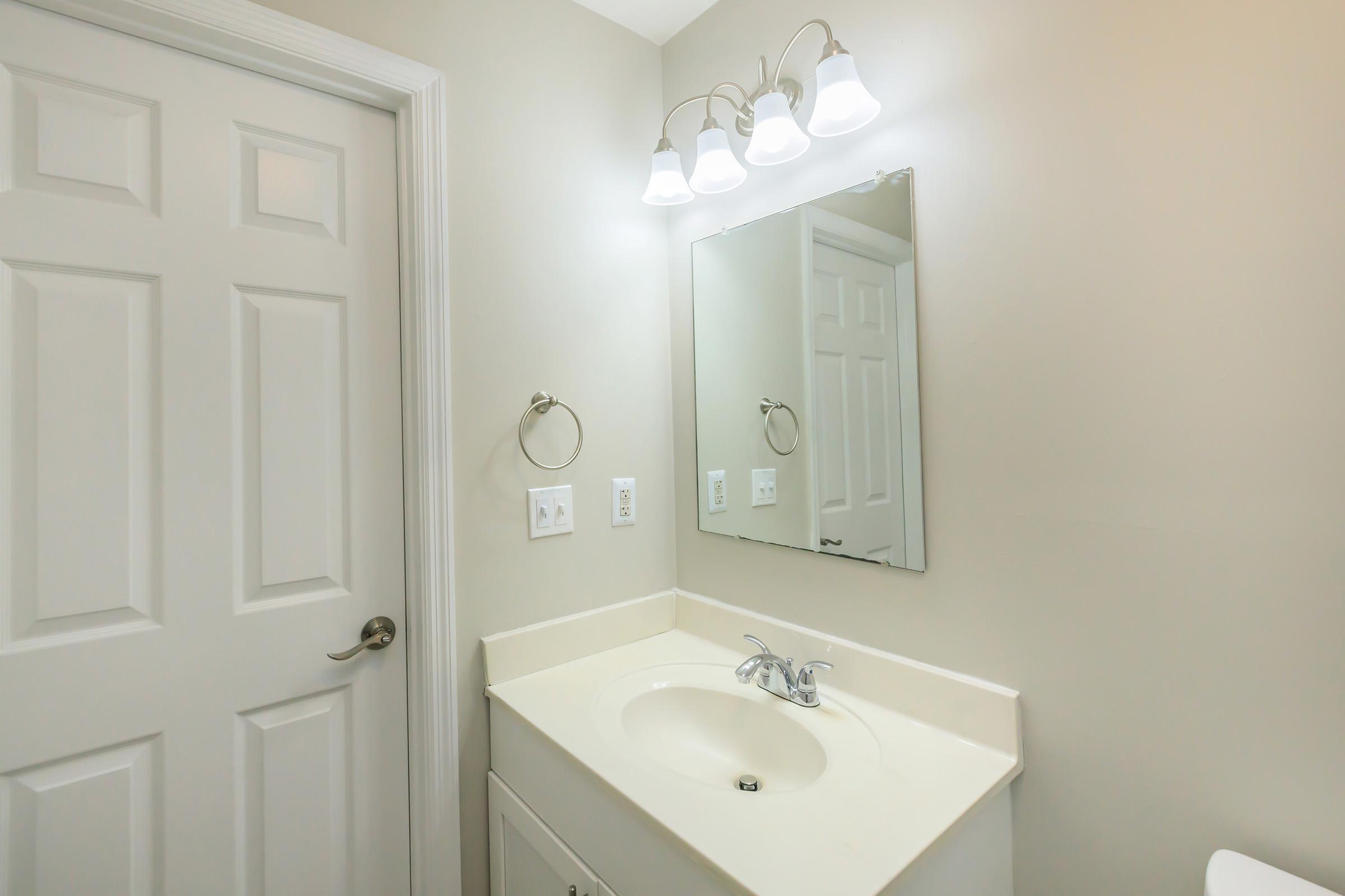 A clean and modern bathroom featuring a white vanity with a sink, a large mirror, and a well-lit area with three light fixtures. A closed white door is visible, and the walls are painted in a neutral beige color, creating a bright and airy atmosphere.