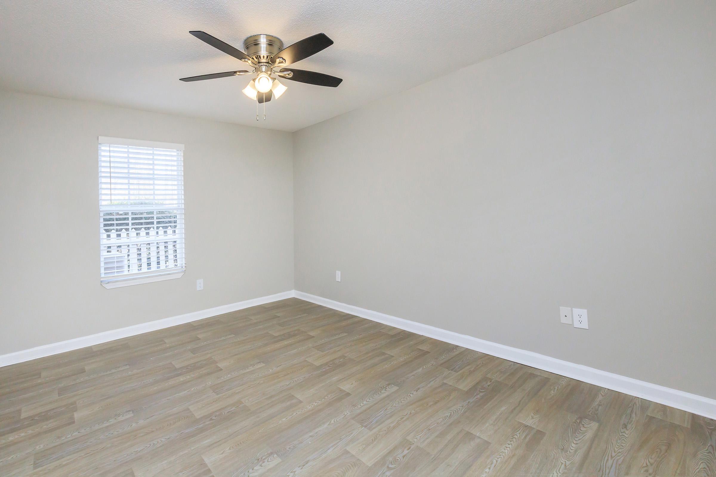 A spacious, empty room with light gray walls and a wood-like laminate floor. There is a ceiling fan with black blades and a small window with white blinds, allowing natural light to enter. The room has a minimalist design, making it suitable for various interior styles.