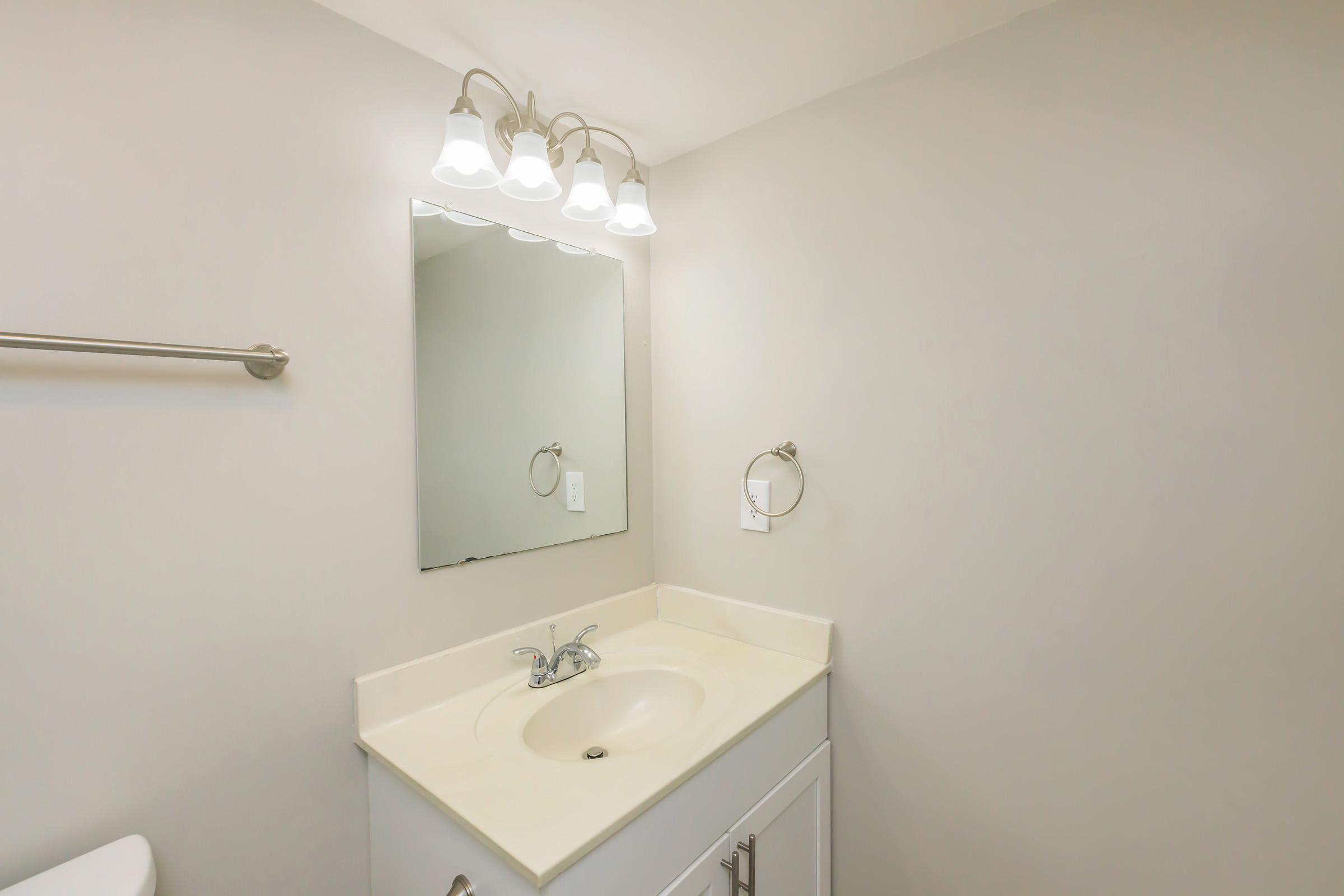 A clean and simple bathroom with a light-colored wall. There is a white vanity with a sink and a faucet. Above the sink, a rectangular mirror reflects the space, and three light fixtures are mounted on the wall. A towel bar and a small towel holder are visible, contributing to the minimalist design.