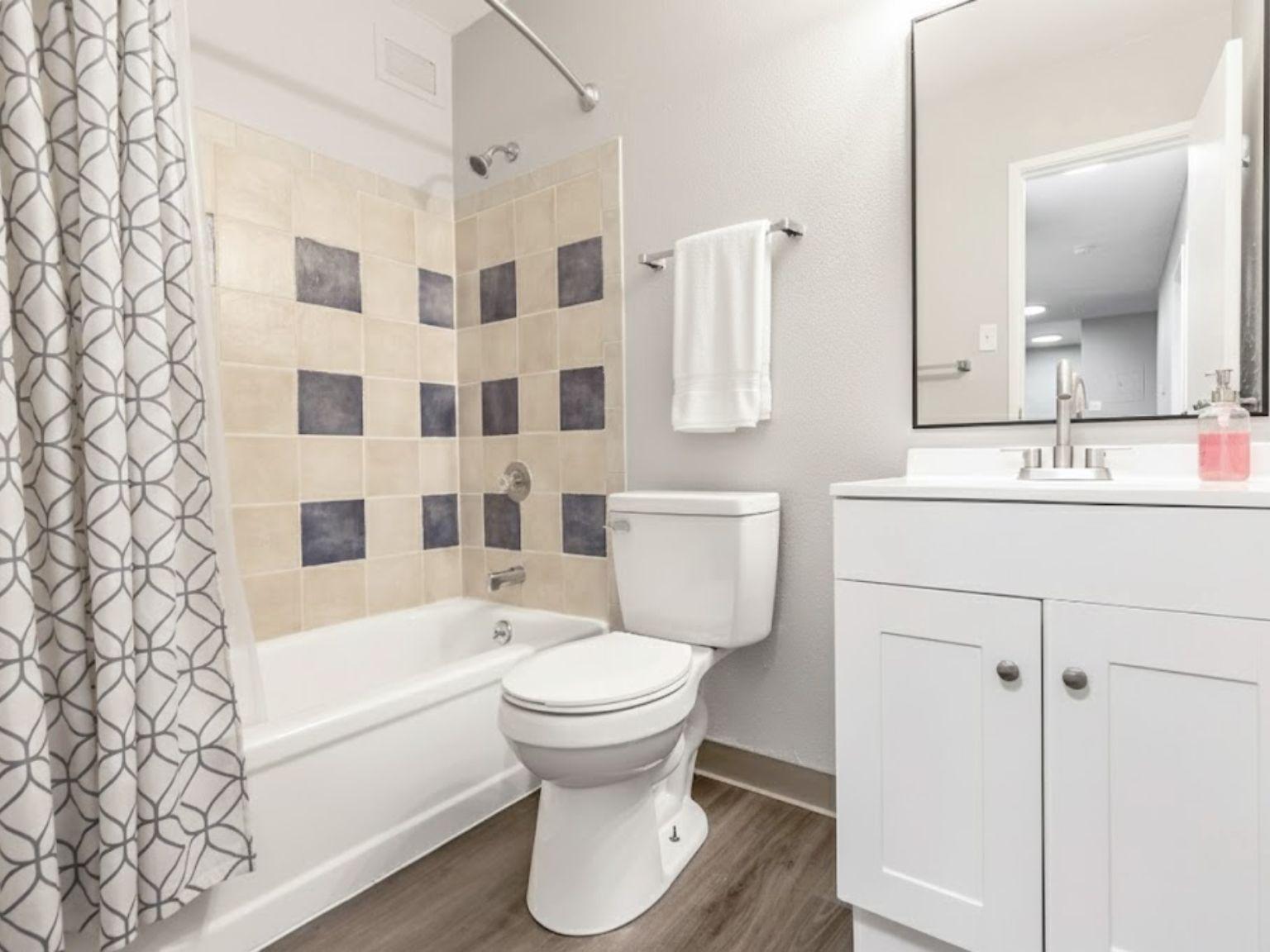 A modern bathroom featuring a white bathtub with a tiled backsplash, a white toilet, and a vanity with a sink. A patterned shower curtain hangs to one side, and there is a white towel rack with a towel. The flooring is a wood-like design, and the space is well-lit with a mirror above the sink.