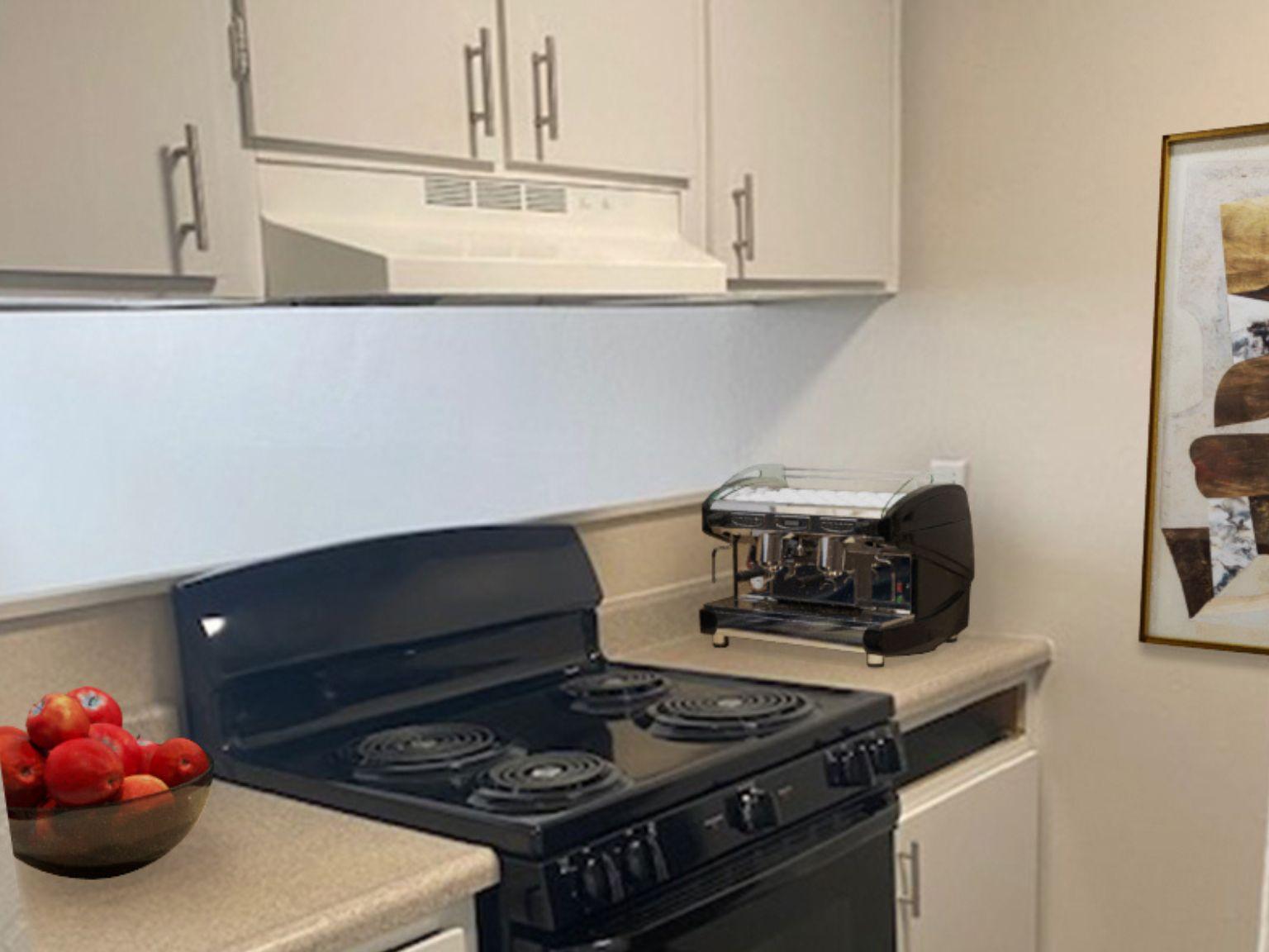 A modern kitchen with light-colored cabinets and a black stove. A coffee machine is placed on the countertop beside a bowl of red apples. There is a range hood above the stove, and a framed abstract artwork hangs on the wall in the background. The overall decor is simple and functional.
