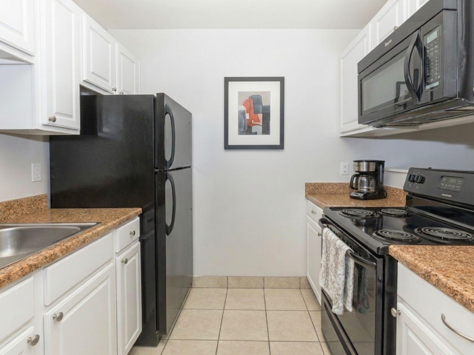 Modern kitchen with white cabinetry, a black refrigerator, and a stainless steel microwave above a black stove. The countertops are a light brown granite. A sink is visible on the left, and a coffee maker is placed near the stove. A framed abstract artwork hangs on the wall, adding a decorative touch. Tile floor is visible.