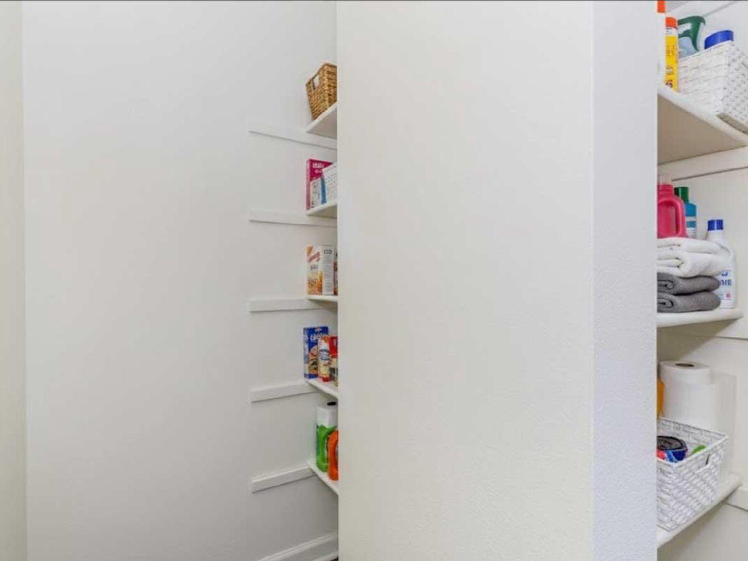 A neatly organized pantry with white walls and shelves. The shelves contain various food items, cleaning supplies, and towels. Some baskets are also present for storage, creating a tidy and functional space.