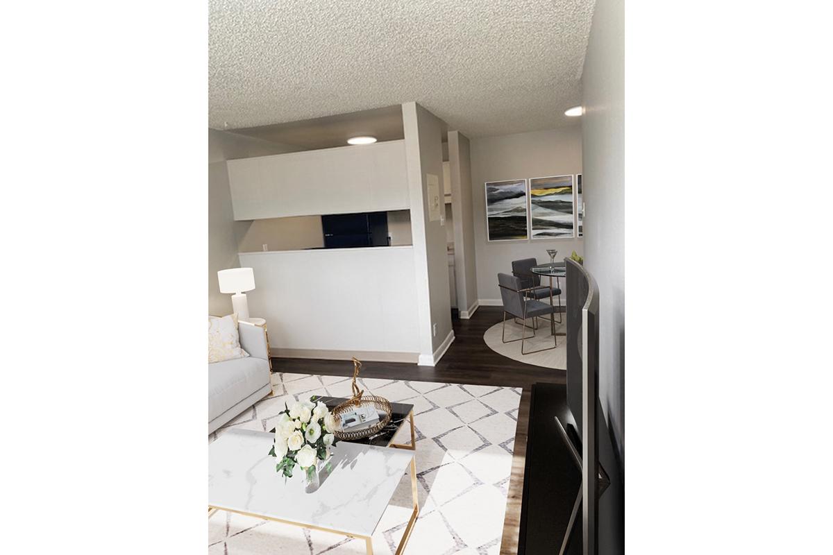 A modern living room featuring a light-colored sofa, a coffee table with a floral arrangement, and a large television. In the background, there is a dining area with a blue wall, a table with a chair, and abstract artwork on the walls. The space has neutral tones and soft lighting, creating a welcoming atmosphere.