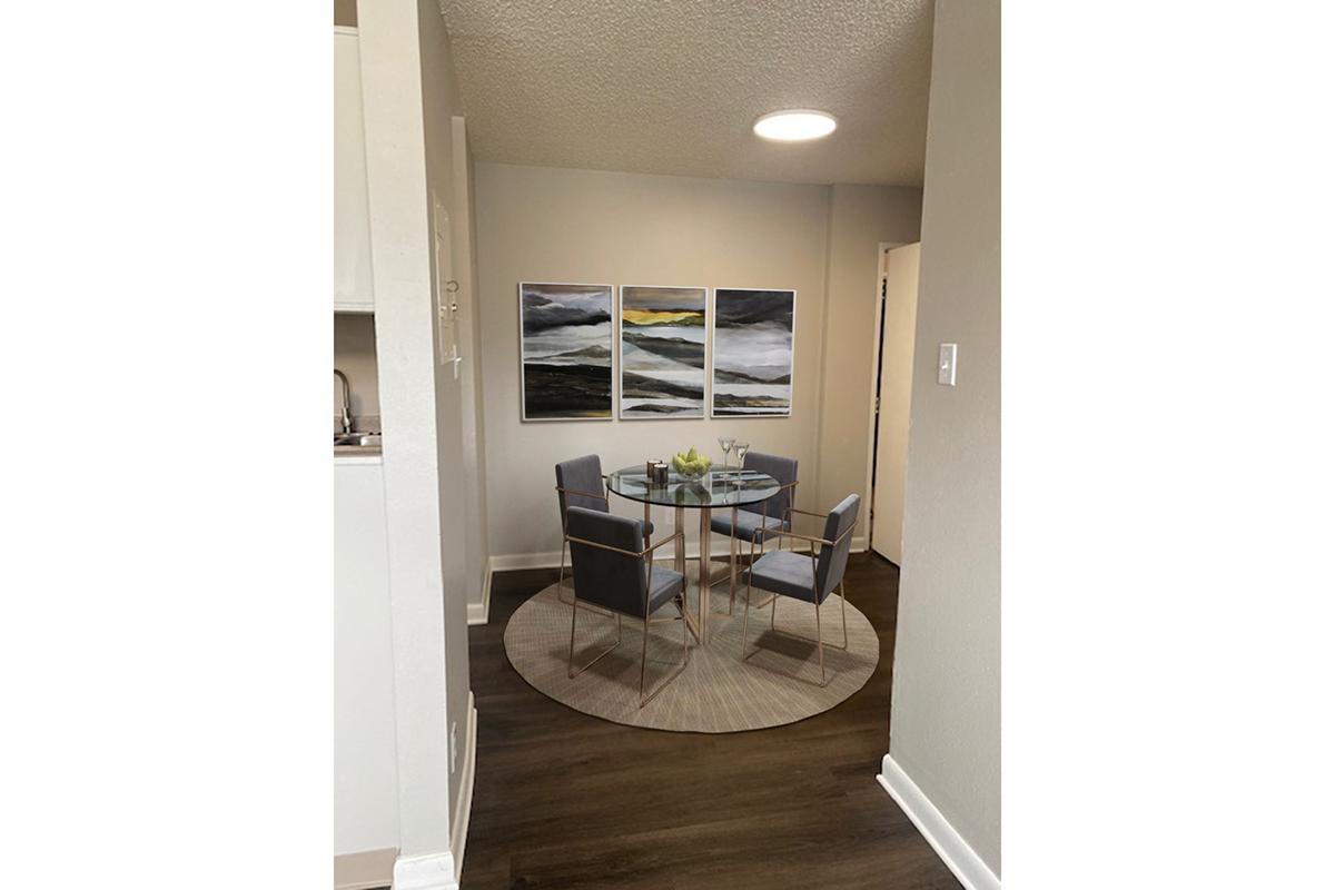 A modern dining area featuring a round glass table with four gray chairs. Above the table, there are three abstract paintings with dark and light gray tones. The walls are neutral-colored, and the floor has a dark wood finish. A round area rug is placed under the table, adding warmth to the space.