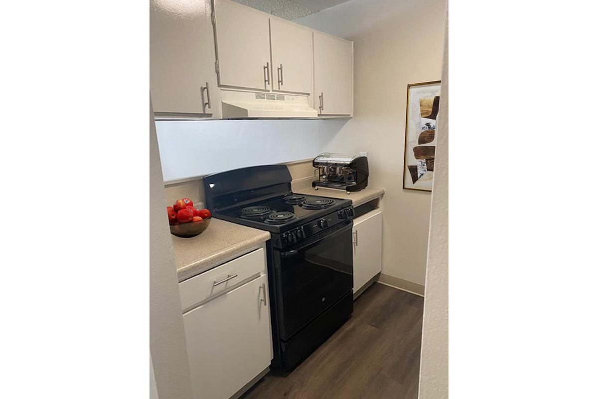 A modern kitchen featuring white cabinets and a black stove. The countertop is light beige, and there's a bowl of red tomatoes on the countertop. A coffee maker is visible next to the stove. The kitchen has an open layout with light-colored flooring and a decorative framed artwork on the wall.