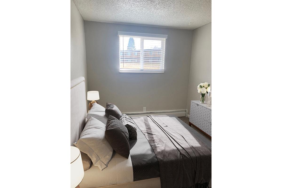A cozy bedroom featuring a neatly made bed with gray and white linens, a bedside lamp, and a white dresser. A window with blinds lets in natural light, illuminating the room. A vase of white flowers adds a touch of elegance to the modern decor.