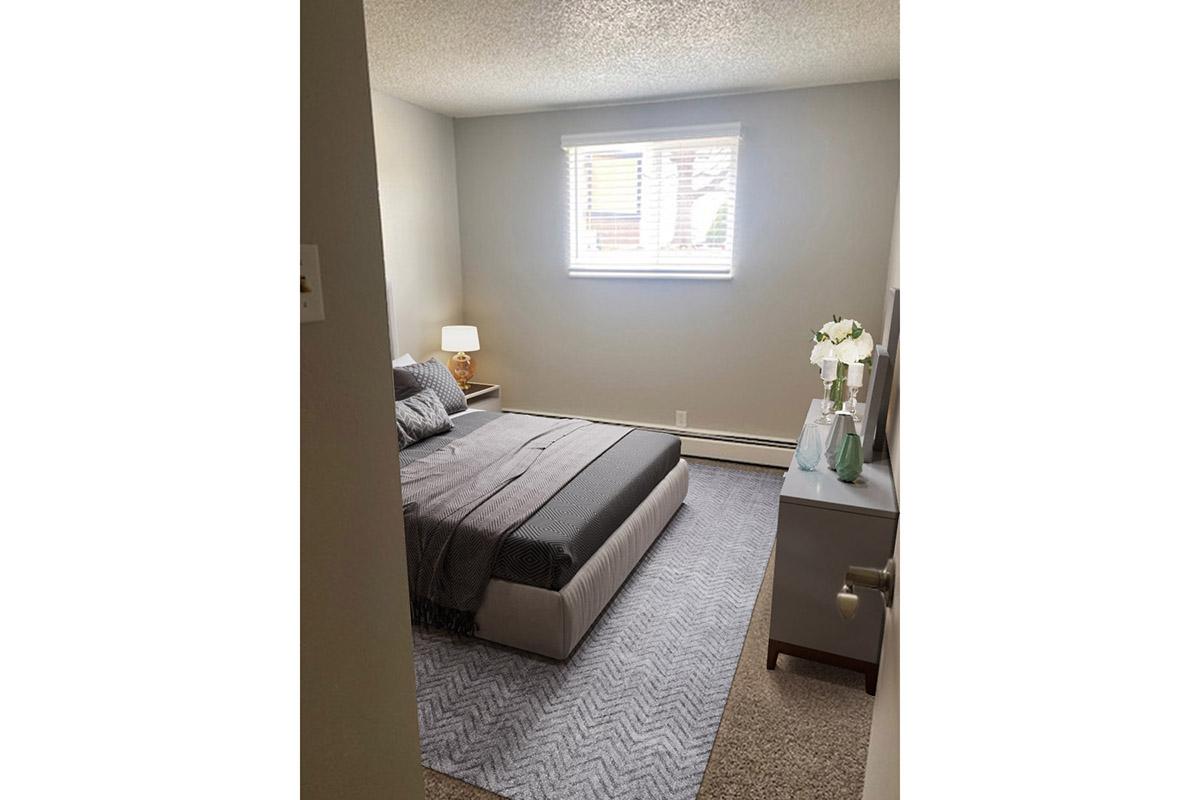 A cozy bedroom featuring a bed with gray bedding, a bedside lamp, and a window with light-filtering shades. There's a small nightstand and a decorative vase with flowers on a dresser against the wall. The room has neutral-colored walls and a soft rug on the floor.