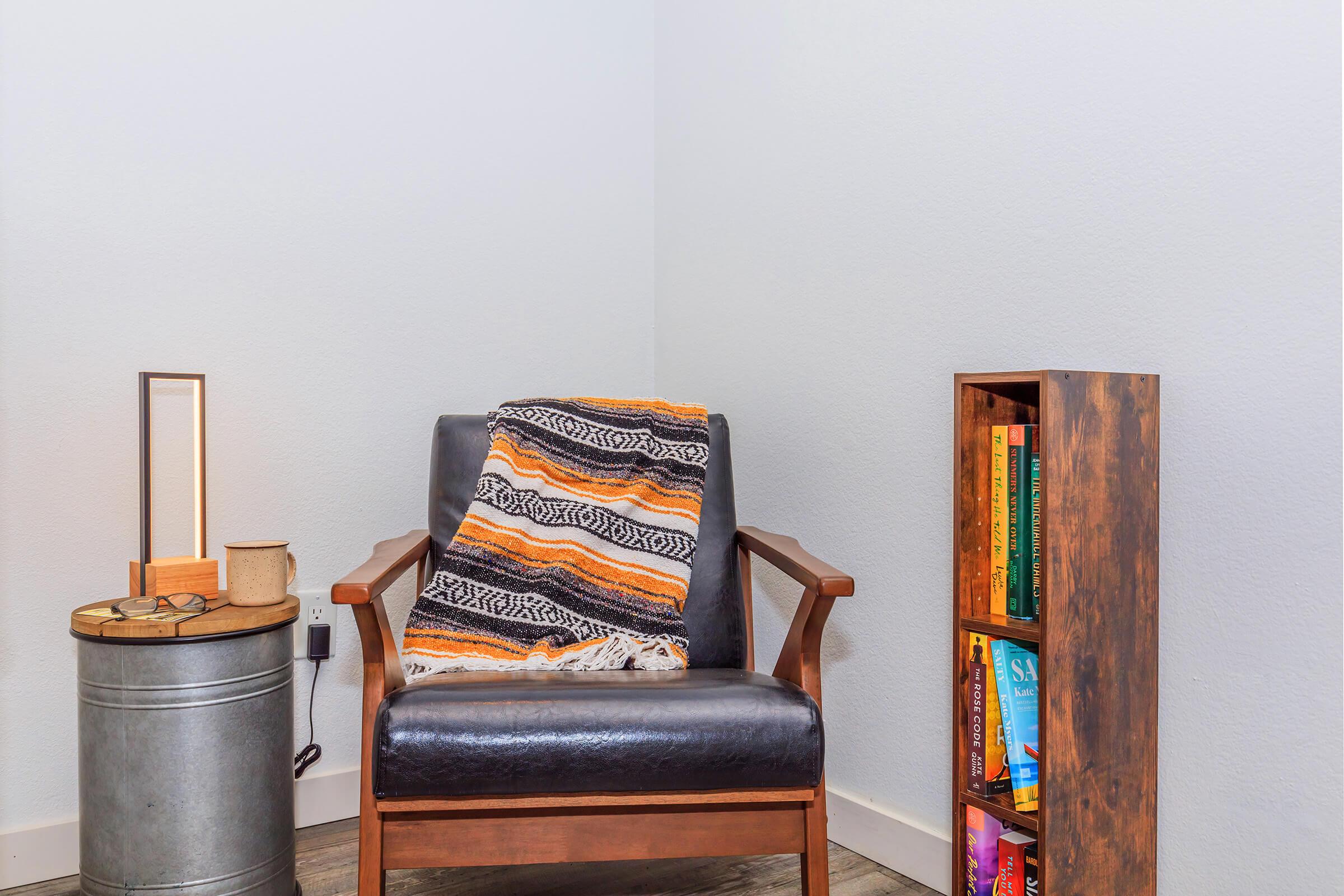 A cozy reading nook featuring a stylish black armchair draped with a colorful striped blanket. Next to it, a wooden side table with a lamp and a small container. A nearby metal bucket complements a tall bookshelf filled with various books, set against a light gray wall.