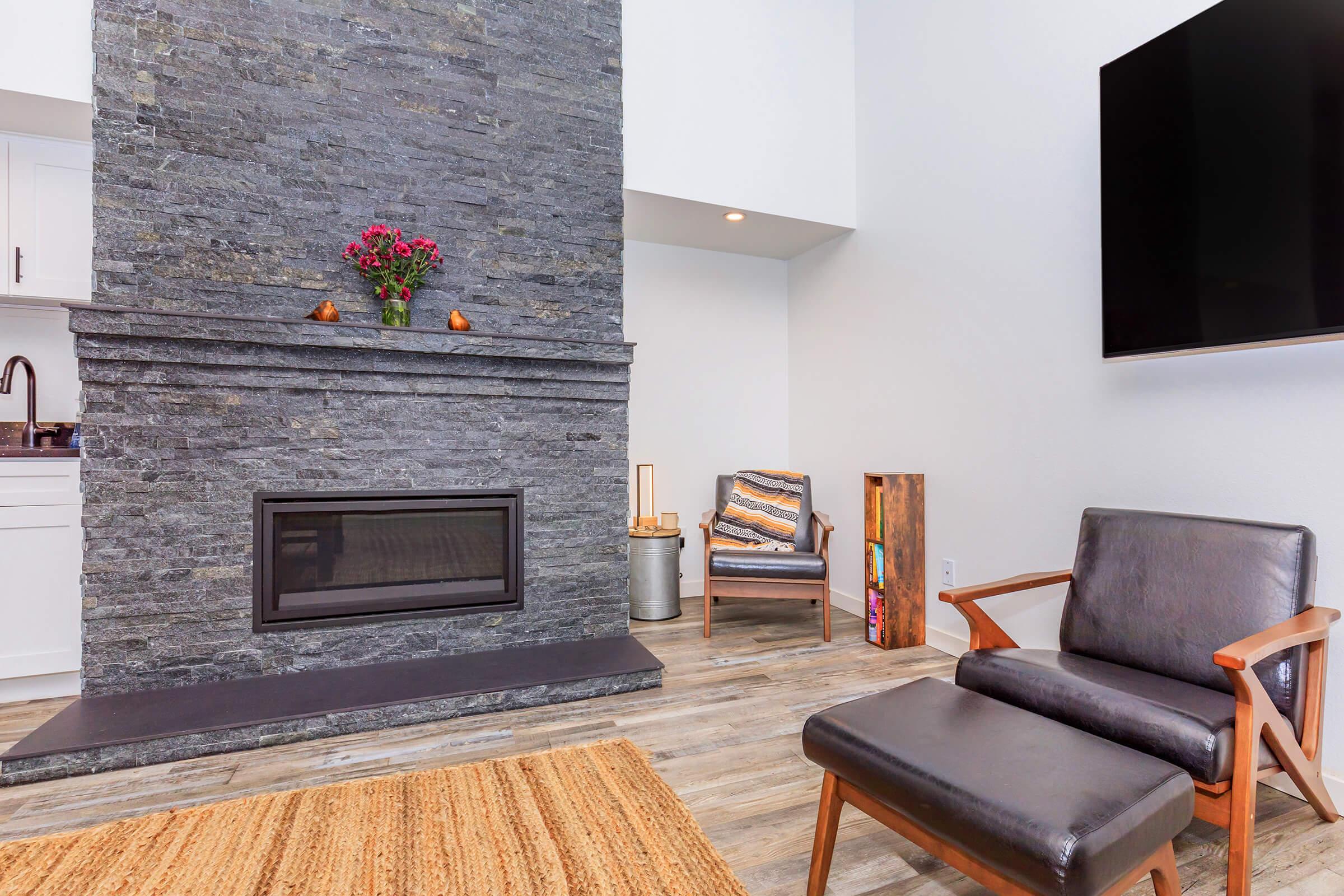 A modern living room featuring a sleek stone fireplace with a flower arrangement on top, a television mounted on the wall, a comfortable black leather chair, and a light-colored rug on the wooden floor. A small side table with decorative items adds to the contemporary aesthetic.