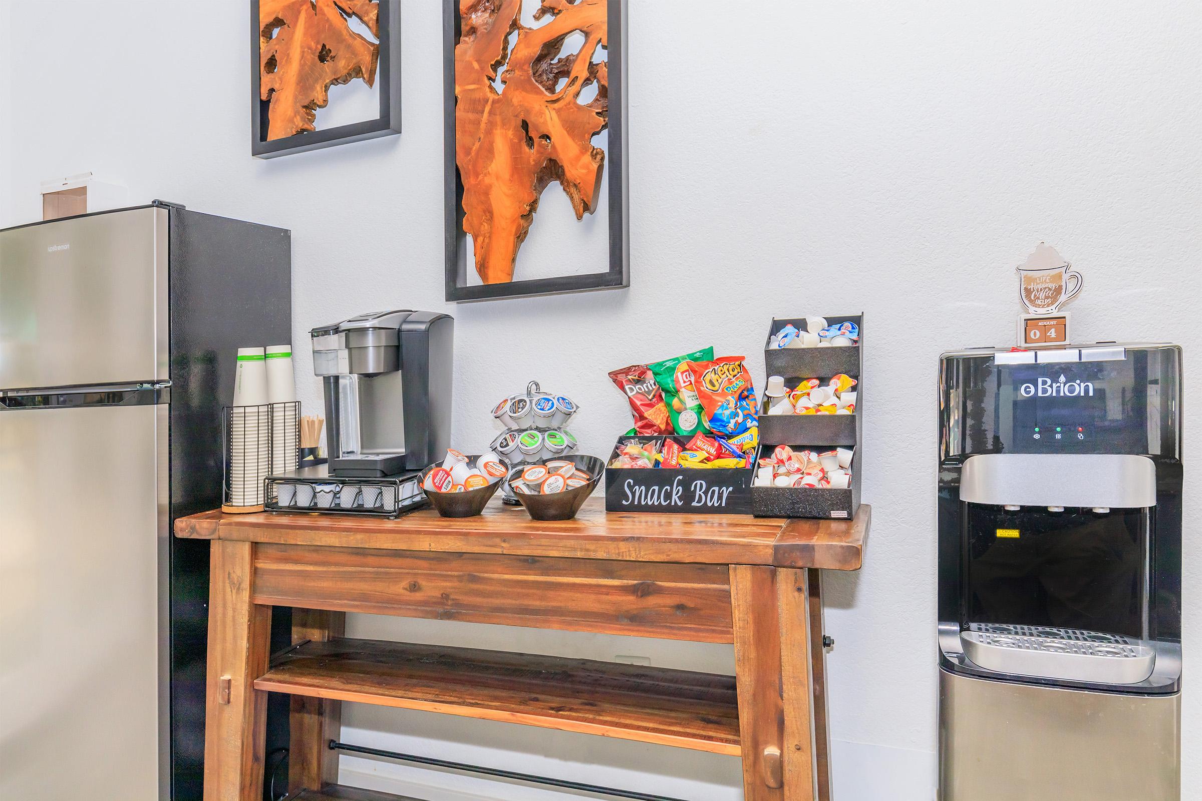 A snack bar setup featuring a wooden table with various snacks in bowls and packages, a coffee machine, a water cooler, and a mini fridge. The wall behind has decorative wooden art pieces. The space is modern and inviting, perfect for a break area.