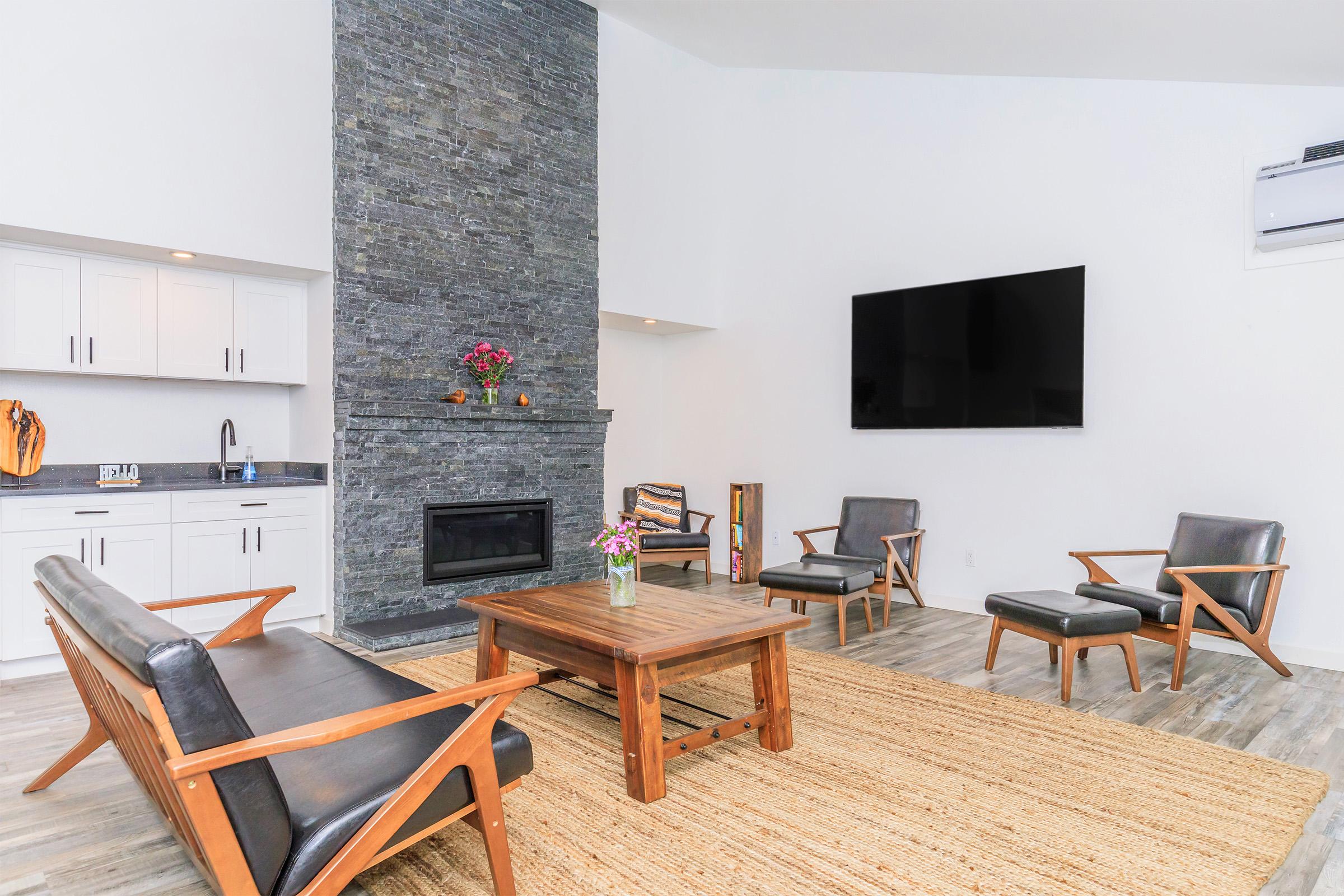A modern living room featuring a stone fireplace, a large flat-screen TV mounted on the wall, and stylish wooden furniture. The seating includes two armchairs and a sofa, arranged around a wooden coffee table. A textured area rug covers the floor, complemented by a vase of flowers on the table.