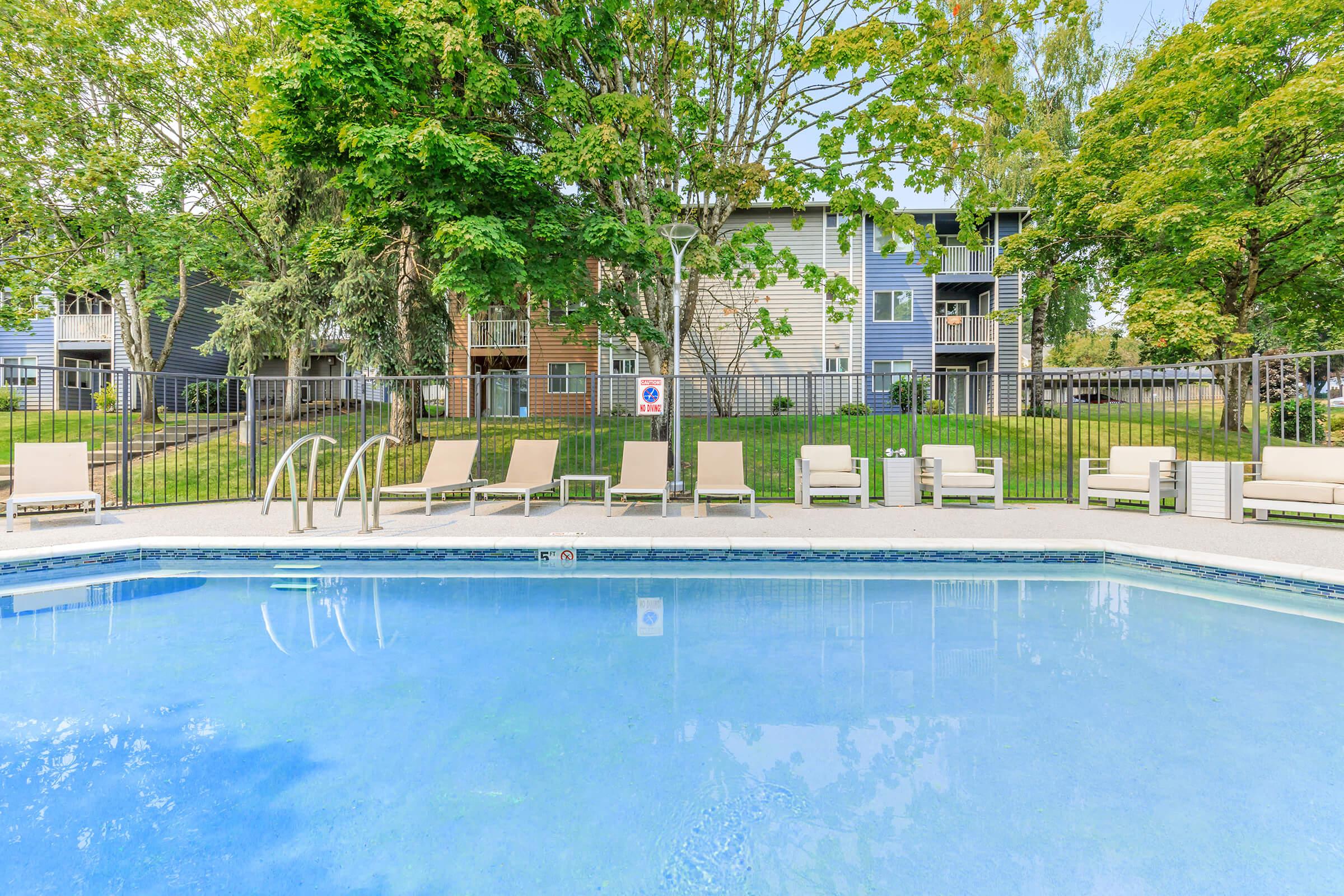 A clear swimming pool surrounded by lounge chairs, set in a well-maintained area with green trees and a residential building in the background. The scene conveys a relaxing outdoor space ideal for leisure and enjoyment.