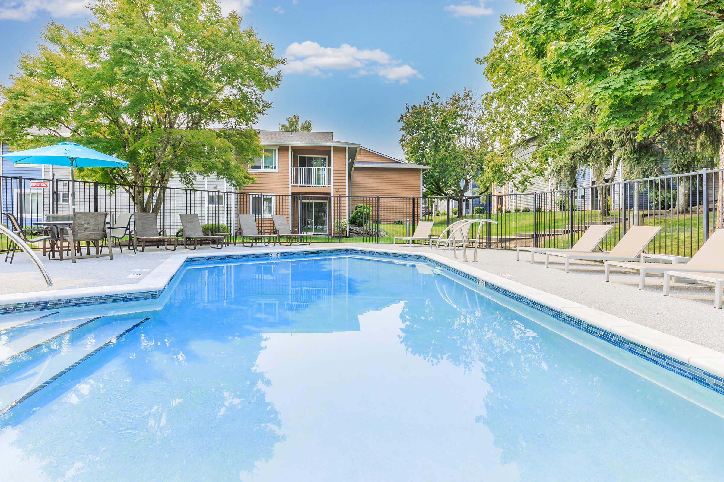 A clear swimming pool surrounded by lounge chairs and an umbrella, set in a landscaped area with green trees. In the background, there are residential buildings with large windows, indicating a relaxing outdoor space for residents and guests.