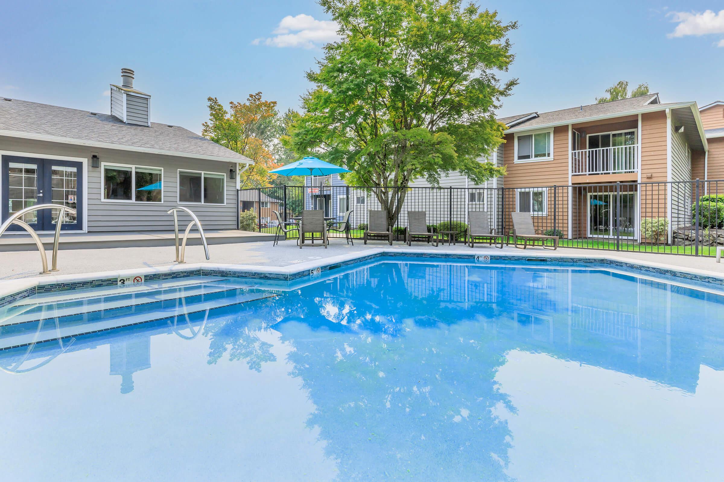 A clear, blue swimming pool surrounded by a patio area with lounge chairs. In the background, a residential building features a balcony and a green lawn. A large tree provides shade, and a colorful umbrella stands near the pool, creating a relaxing outdoor atmosphere.