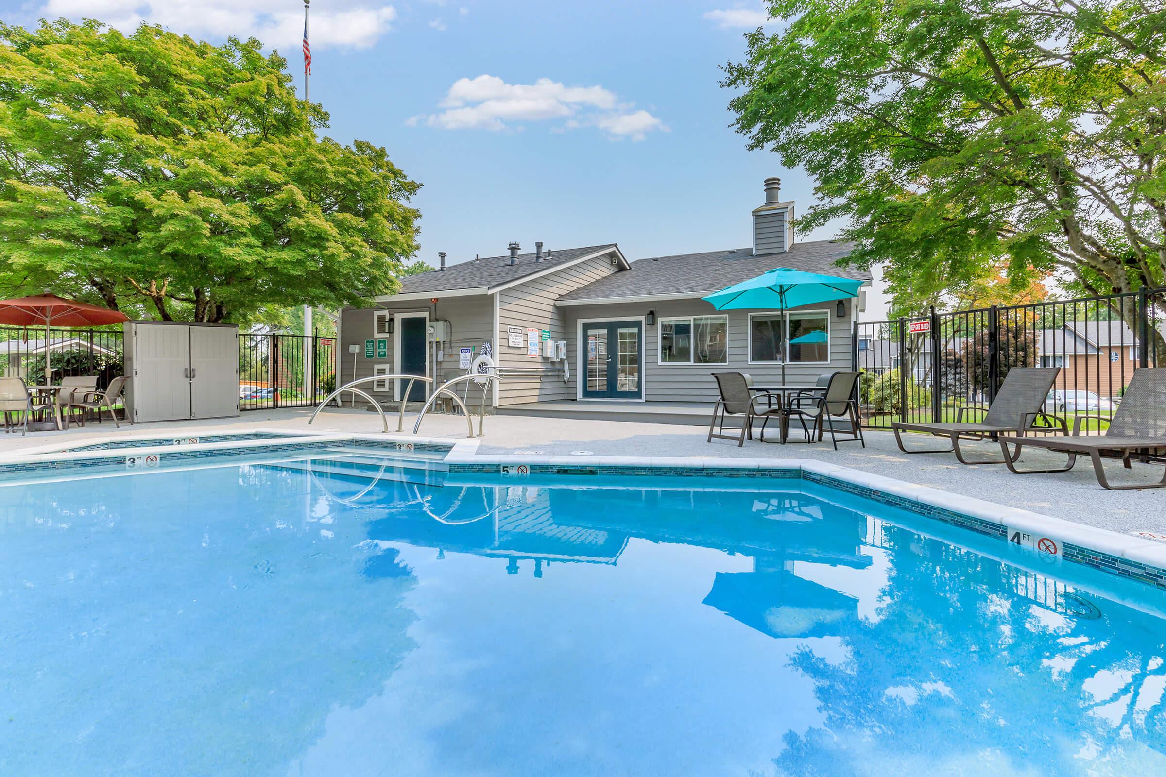 A bright outdoor pool area with clear water reflecting the sky. Surrounding the pool are lounge chairs and umbrellas, with a clubhouse in the background. Lush green trees provide shade, and a fence lines the space, creating a relaxing atmosphere for leisure and recreation.