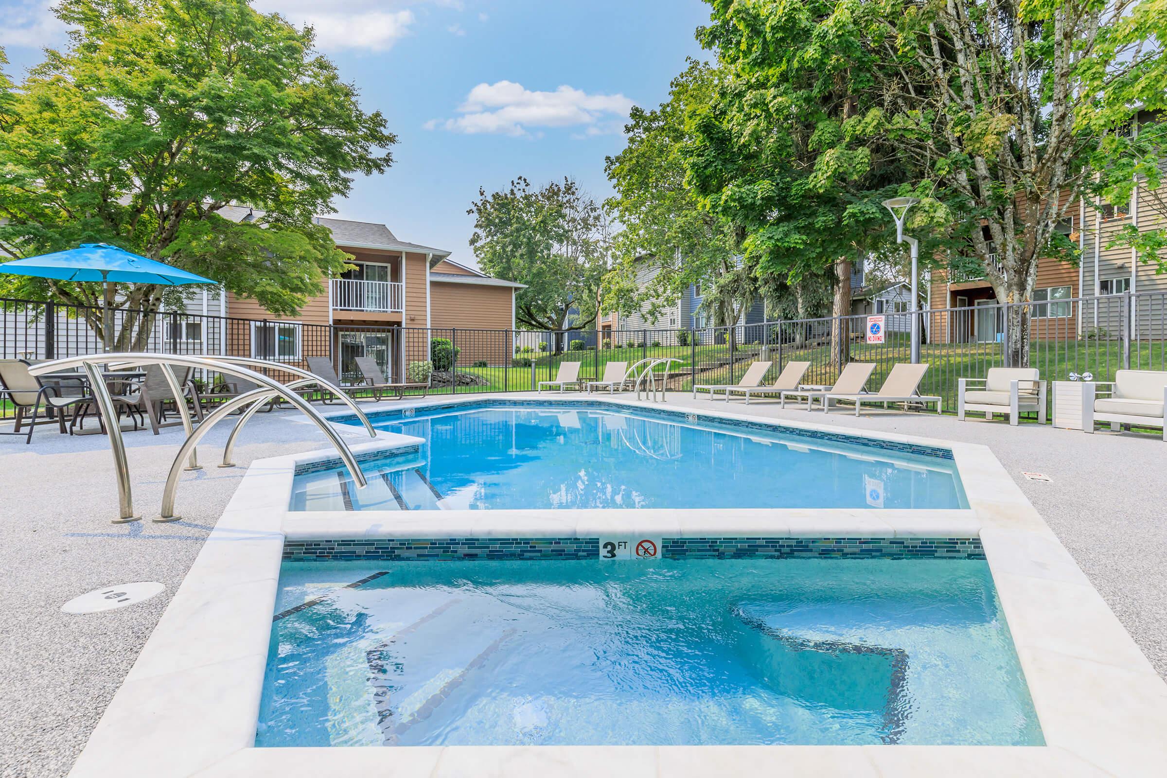 A clear blue swimming pool with a curved edge and stairs, surrounded by lounge chairs and shaded by trees. Nearby, there are residential buildings and a patio area with umbrellas. The pool area is well-maintained and inviting, under a bright blue sky with a few clouds.