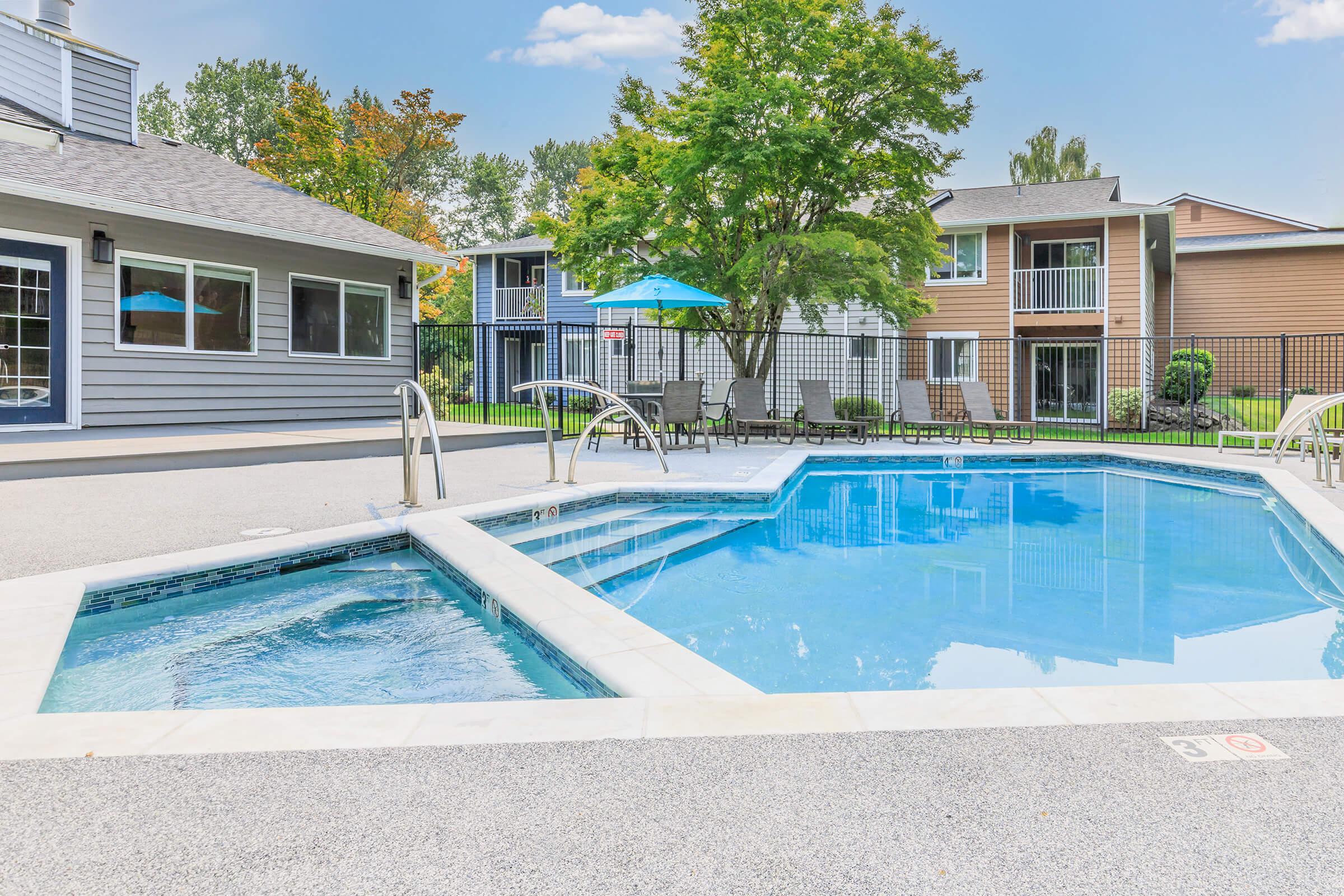 A modern swimming pool featuring a connected hot tub, surrounded by a patio area with lounge chairs and umbrellas. In the background, there are two multi-story residential buildings and lush greenery. The scene is bright and inviting, ideal for relaxation and leisure.